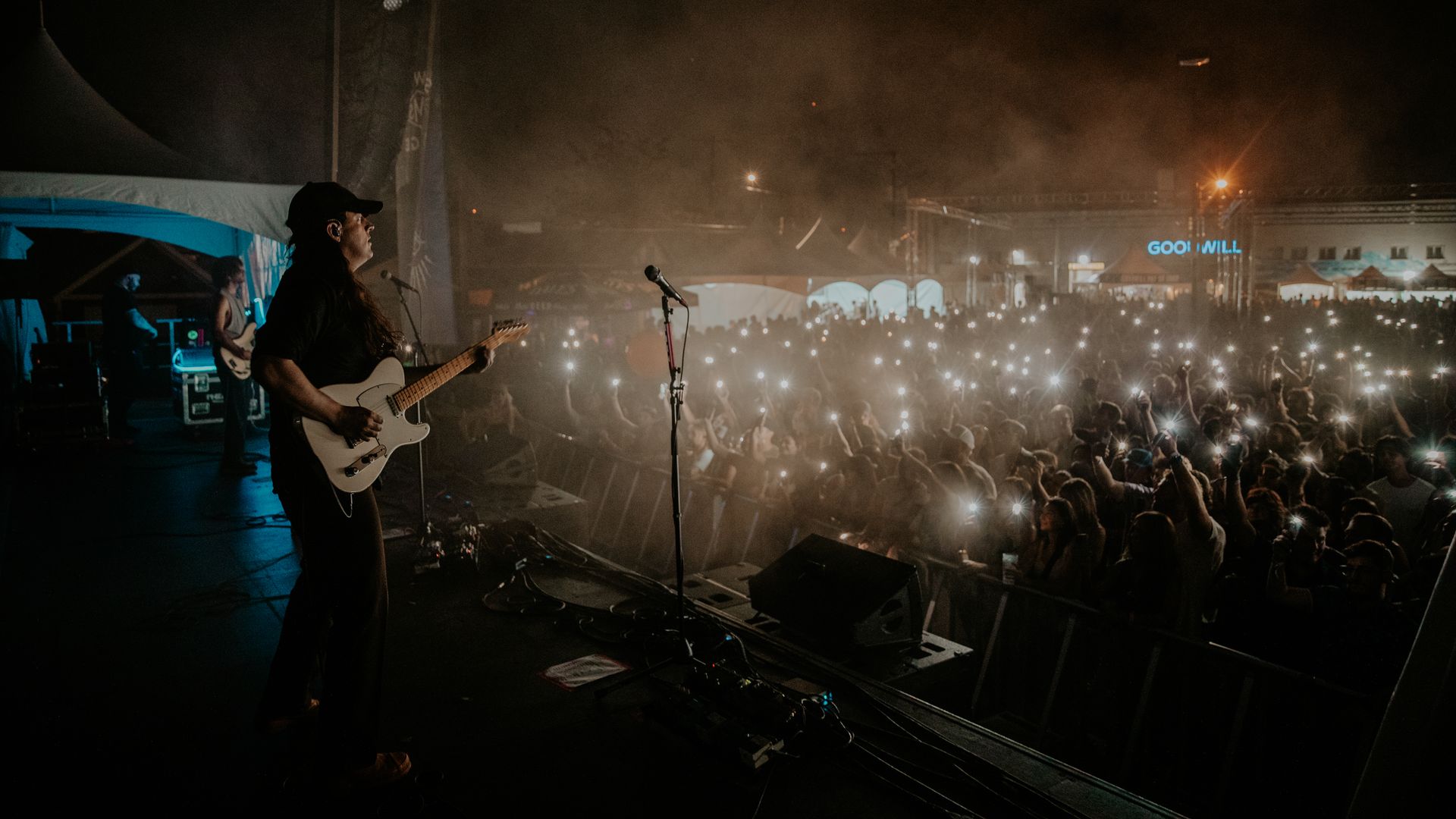 Nighttime concert stage with a guitarist in black and a white electric guitar on the left; a microphone stand nearby. A large crowd raises phones with lights, hazy air, and a distant "Goodwill" sign.