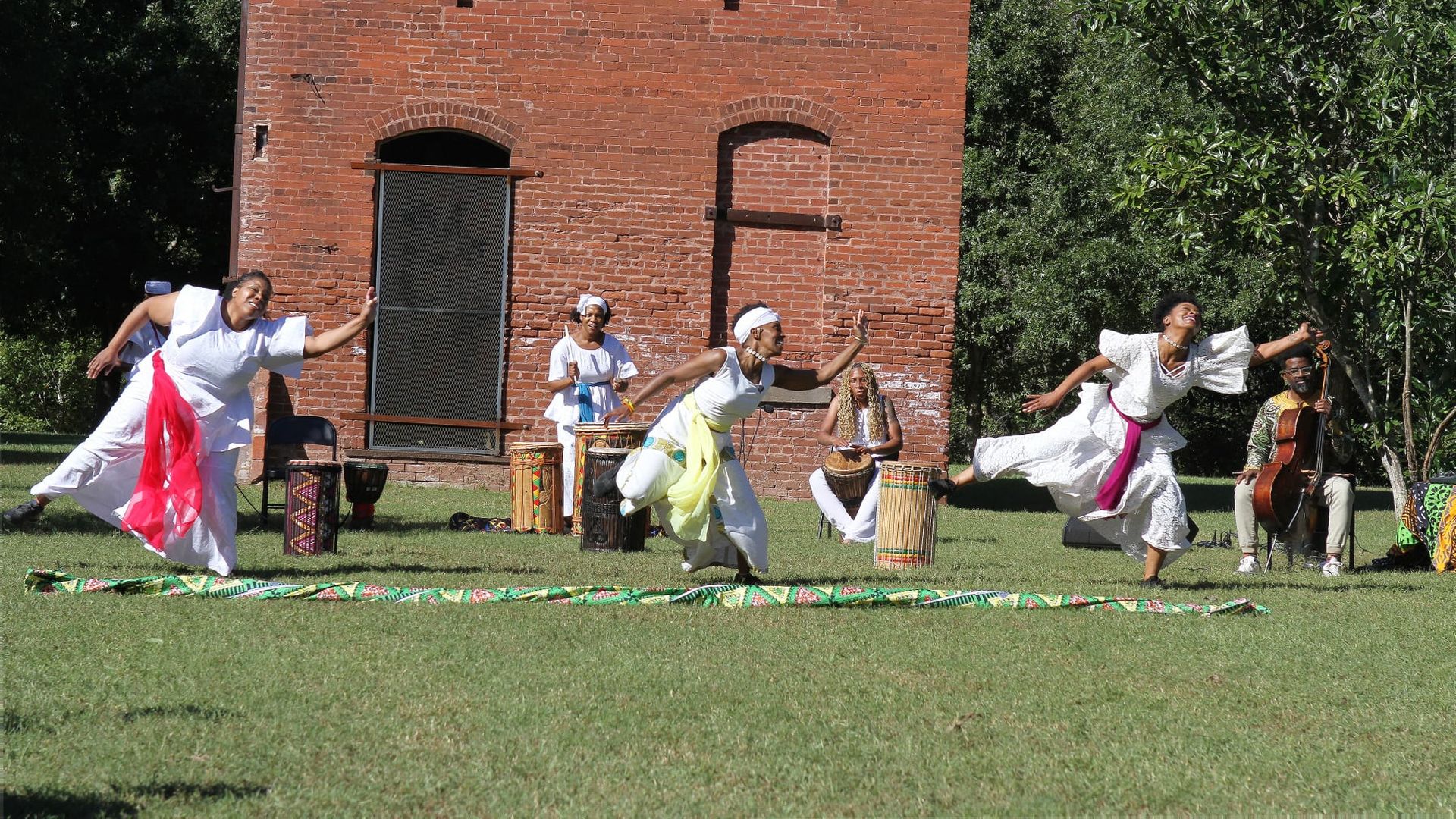 Three women in white dresses performing a dance on grass in front of a red brick building, with drummers and a cellist playing music nearby under trees.