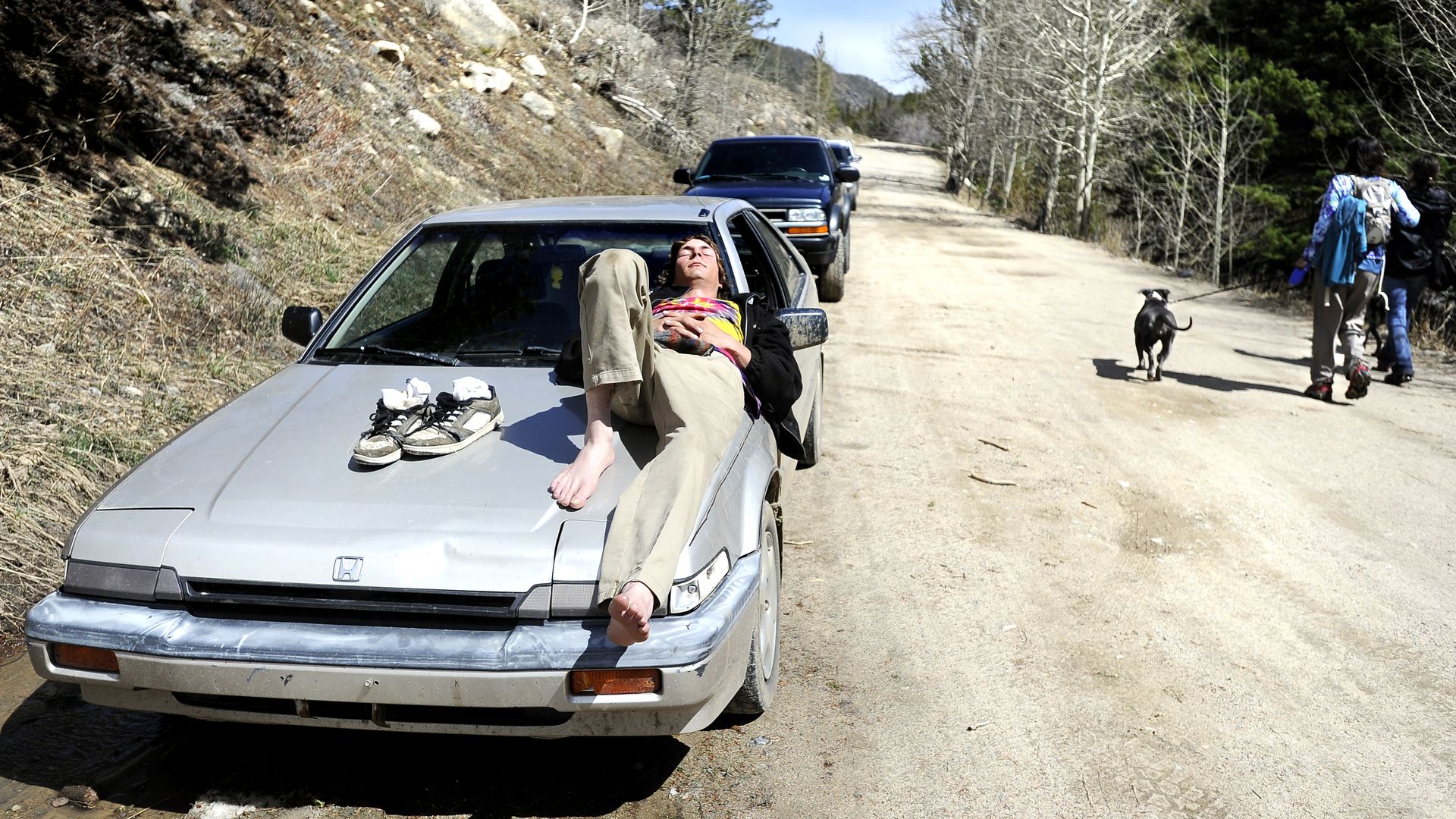 Image of a hiker napping on the hood of his car near a trailhead while hikers walk by. 