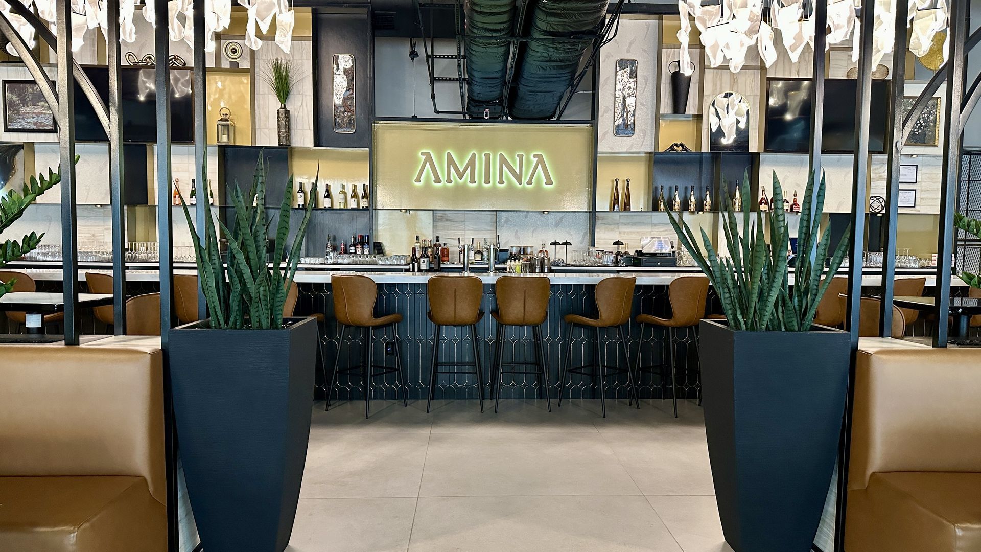 Chic bar scene with a glowing "AMINA" sign above brown leather stools at a blue tiled counter. Two tall planters with snake plants flank beige banquettes as glass shelves and art decorate the space.