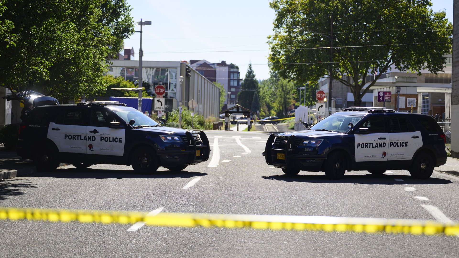 Two Portland Police SUVs block a city street, yellow caution tape in the foreground, trees on both sides, and urban buildings in the background on a sunny day.