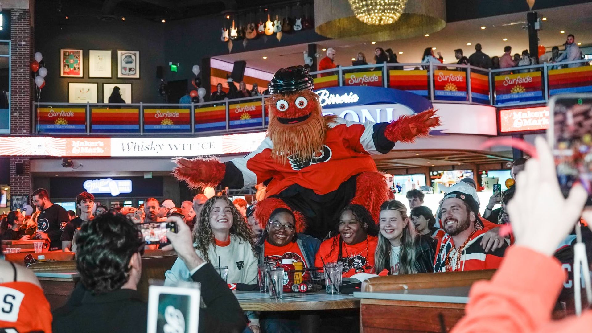Crowded bar scene with the Flyers mascot, Gritty, wearing a black helmet, lifted above cheering fans.