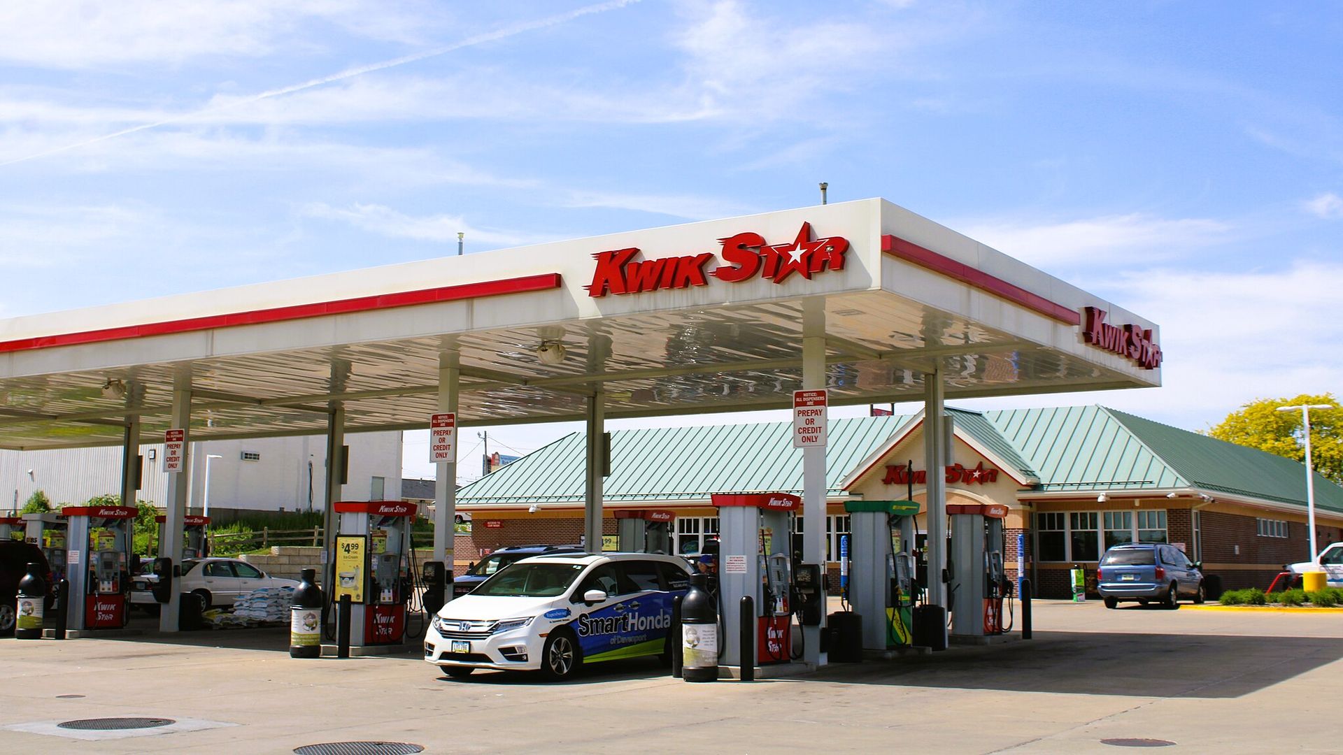 Kwik Star gas station with red signage, multiple fuel pumps, a white car with SmartHonda branding, and a building with green roof under a blue sky with light clouds.