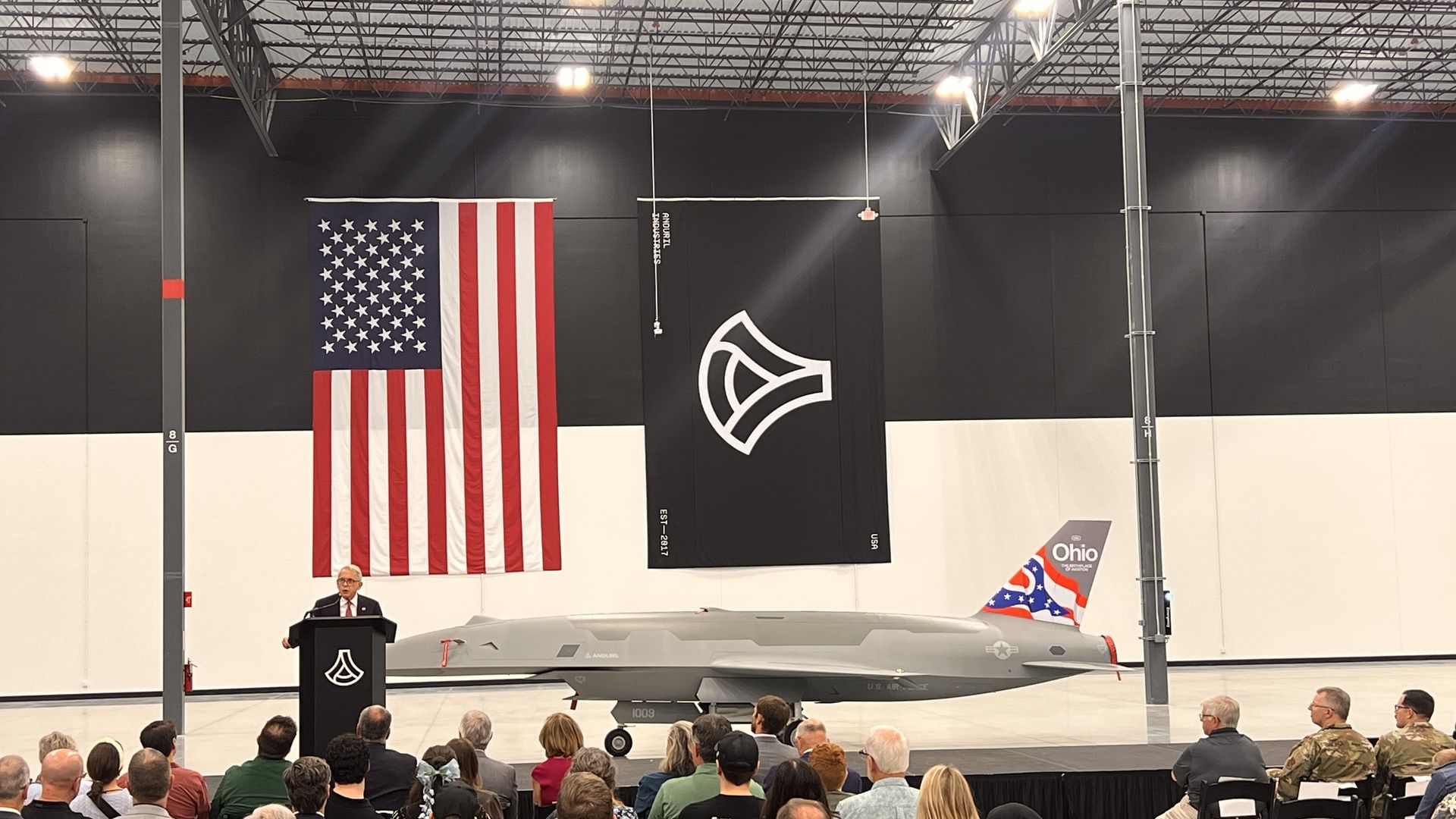 Indoor hangar with a gray military jet on the floor, a speaker at a podium, and a large American flag hanging beside a black banner with a white emblem; audience seated and watching.