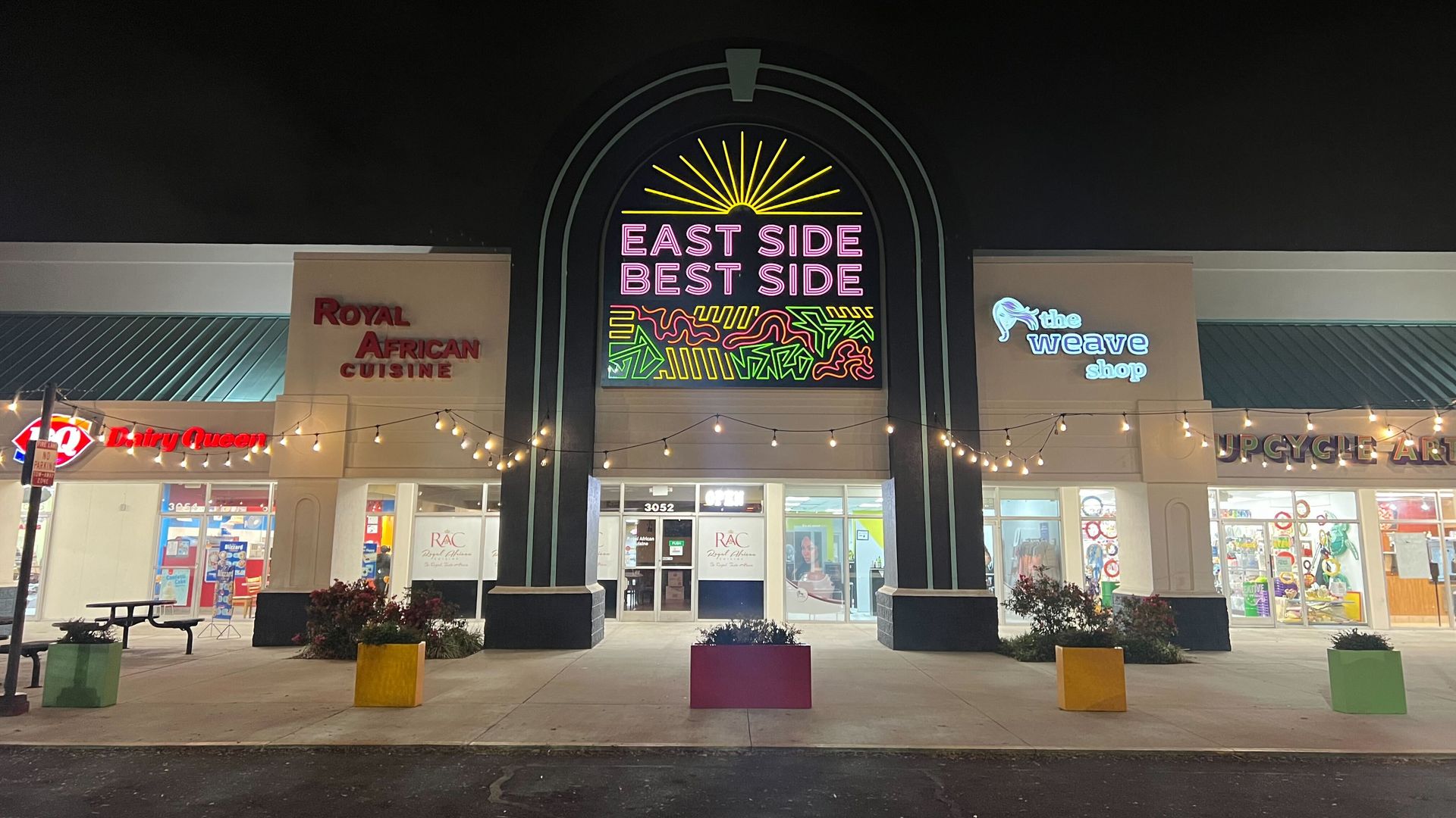 Nighttime storefronts with a central neon sign reading "East Side Best Side" beneath a sunburst arch; signs for "Royal African Cuisine" to the left and "the weave shop" to the right, plus string lights.