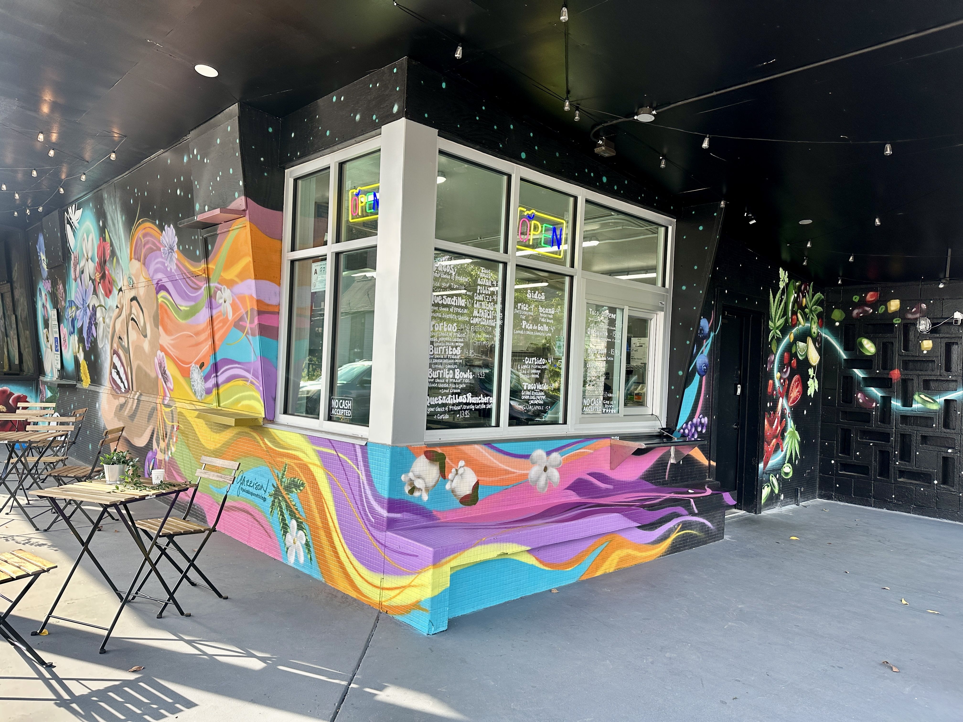 Outdoor cafe with colorful mural of a smiling woman with flowing rainbow hair and flowers. Black ceiling with string lights, open window with neon "OPEN" sign, and tables with chairs.