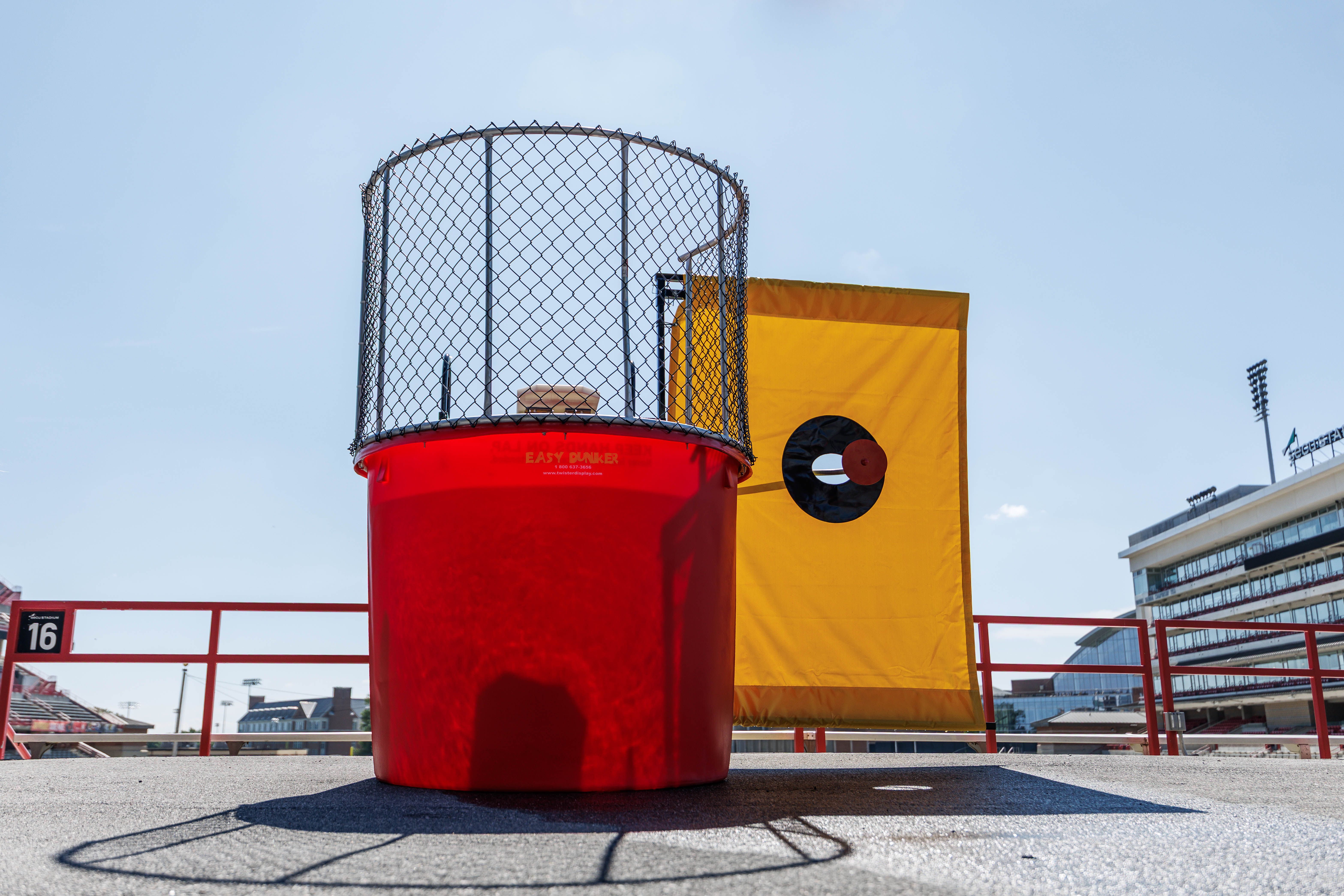 Close-up of a large red cylindrical structure with a black metal chain-link cage on top, beside a yellow banner with a black circle, at an outdoor sports stadium under a blue sky.