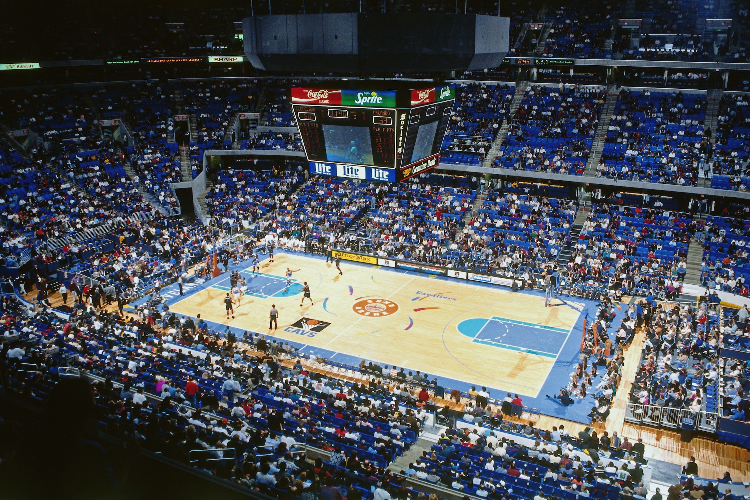 Indoor basketball arena with blue seats; players on a polished court featuring a central logo, while fans fill the stands. A large overhead scoreboard displays scores and ads.