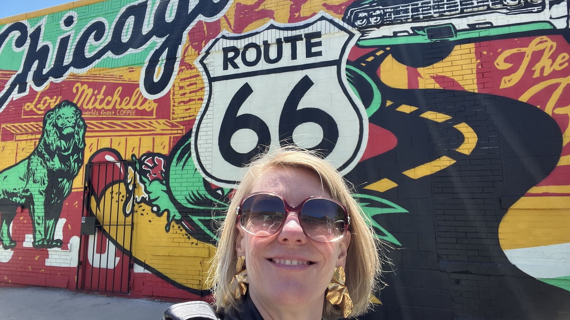Colorful mural on a brick wall featuring a Route 66 shield and Hollywood script, with green, yellow, and red hues. In the foreground, Carrie Shepherd wears oversized sunglasses.
