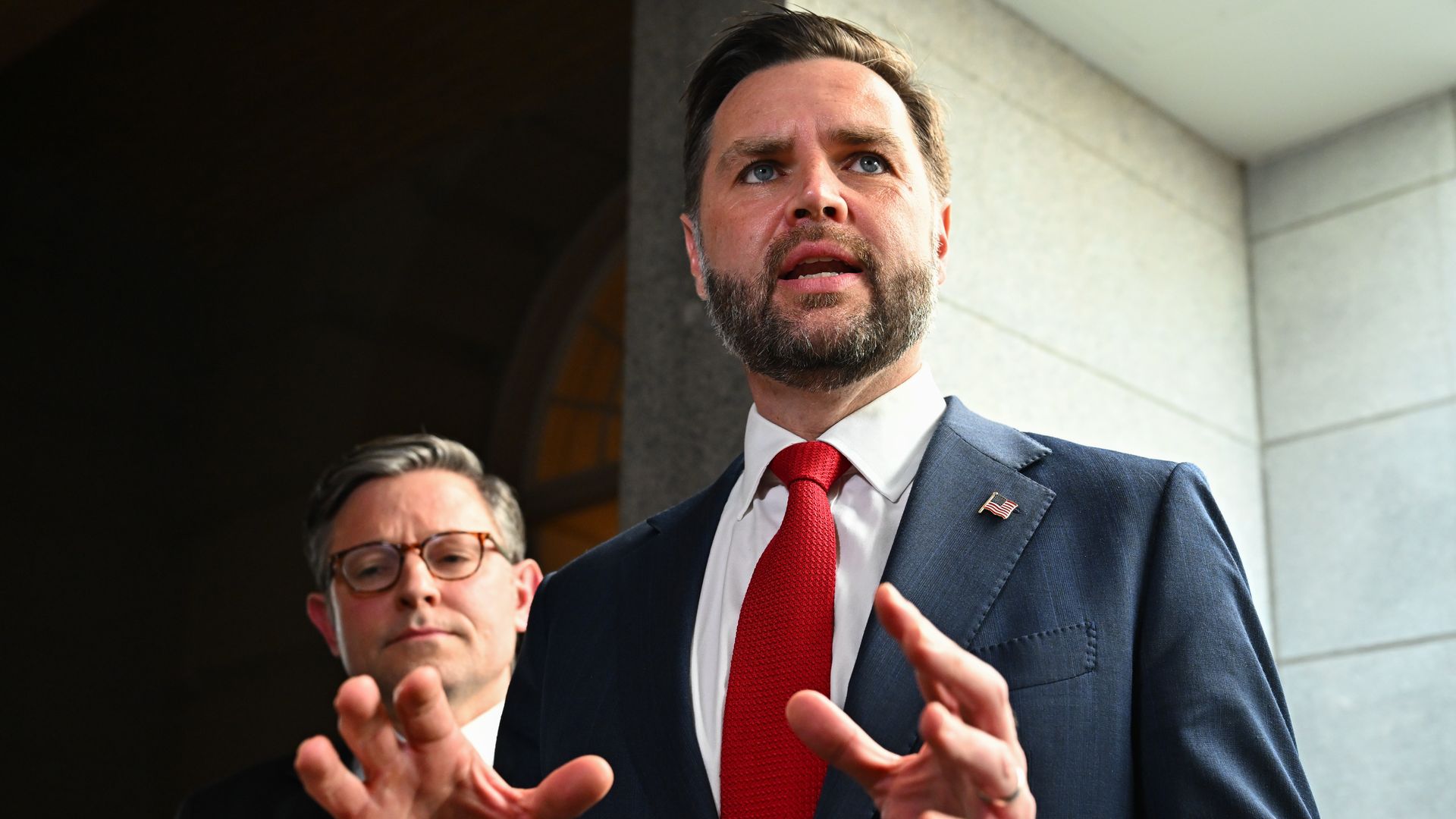 A bearded man in a navy suit, white shirt, and red tie speaks passionately outdoors, with a small American flag pin on his lapel; another man is partly visible in the foreground.