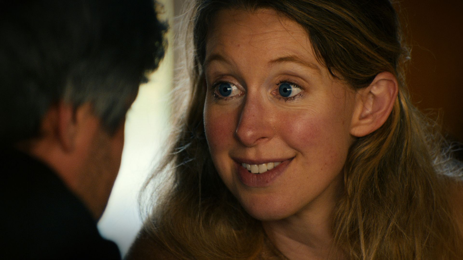 Close-up of a smiling woman with blue eyes and light brown hair, looking toward a person off to the left. Dark background on the left, warm brown lighting on the right.