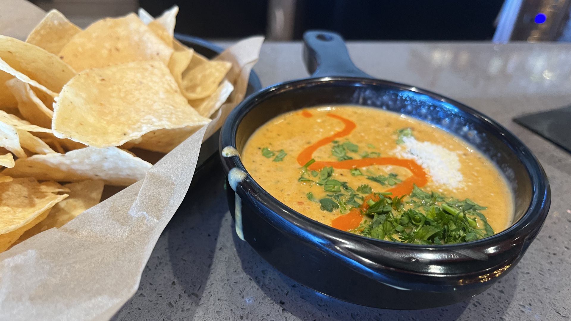 A bowl of queso with cilantro and diablo sauce next to a basket of chips. 