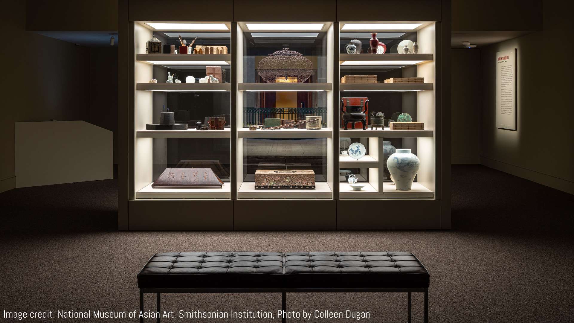 Museum display case with diverse Asian art objects including pottery, wooden artifacts, a large blue and white vase, and a black bench in front of the case under soft lighting.