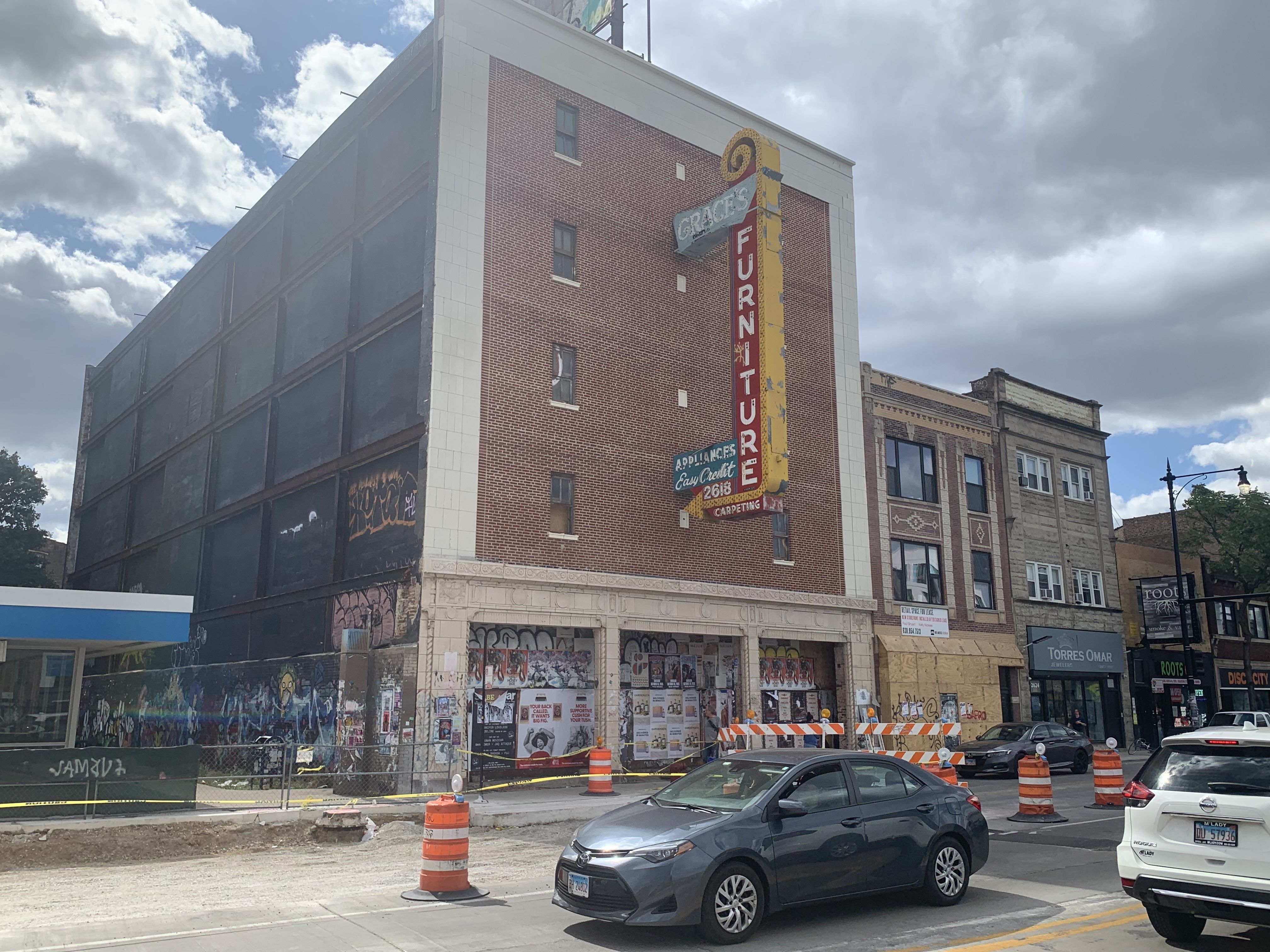 Street view of an old brick building with a large faded red and yellow sign reading "Grace's Furniture." Construction barriers and orange barrels block part of the street; several parked cars are visible.