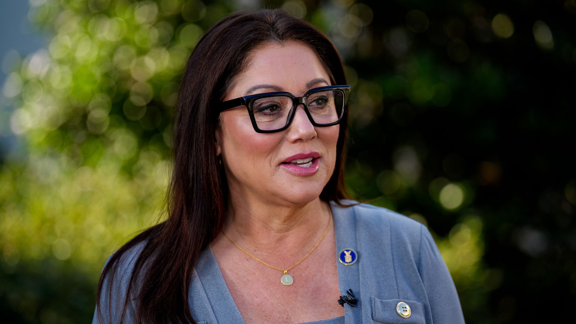 Former Labor Secretary Lori Chavez-DeRemer — a woman with dark hair wearing a gray sweater and top and black glasses — stands and speaks outside near greenery.