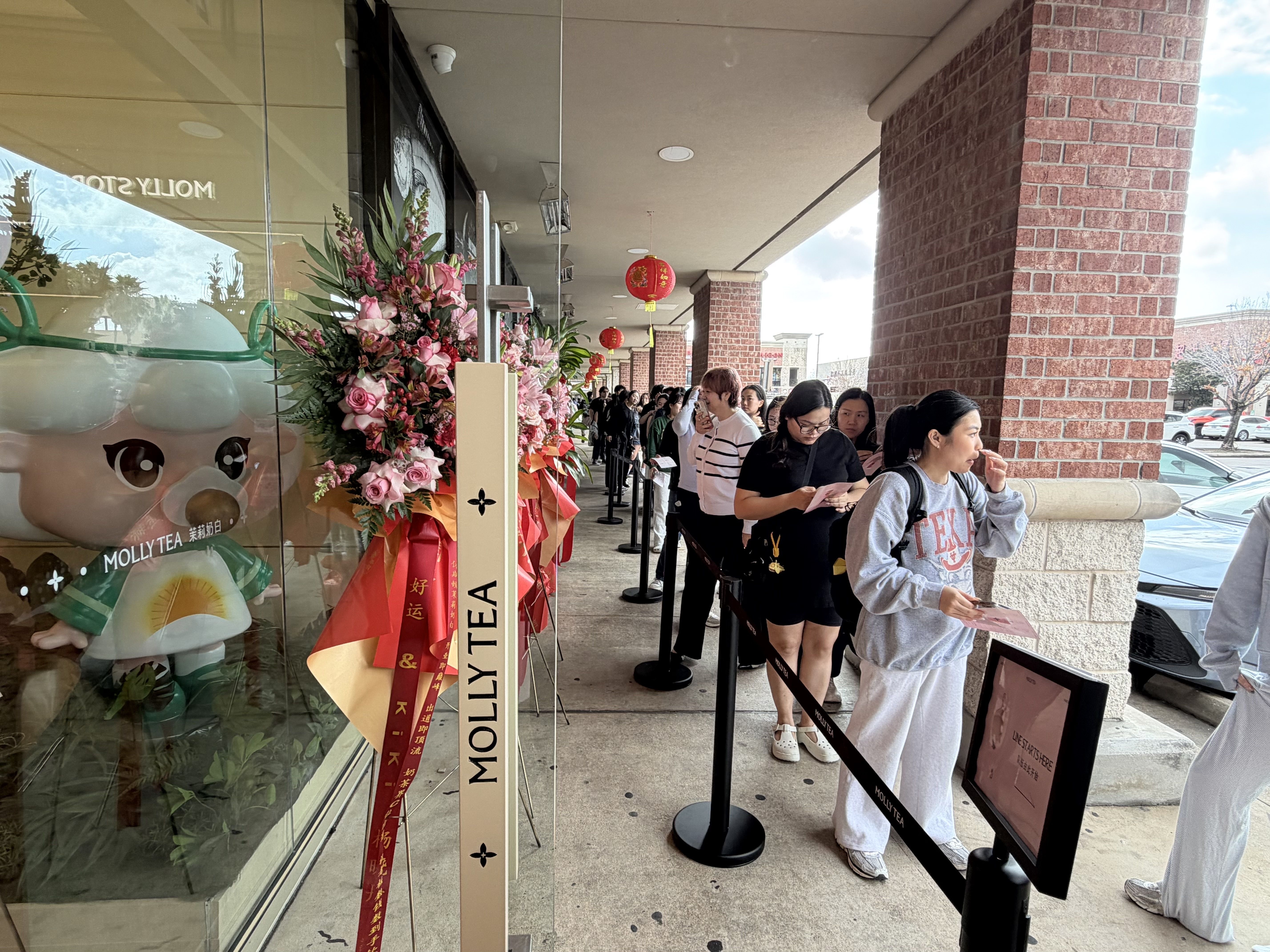 People lined up outside Molly Tea shop, waiting under red Chinese lanterns and next to large pink floral arrangements and a cartoon bubble tea mascot in green.