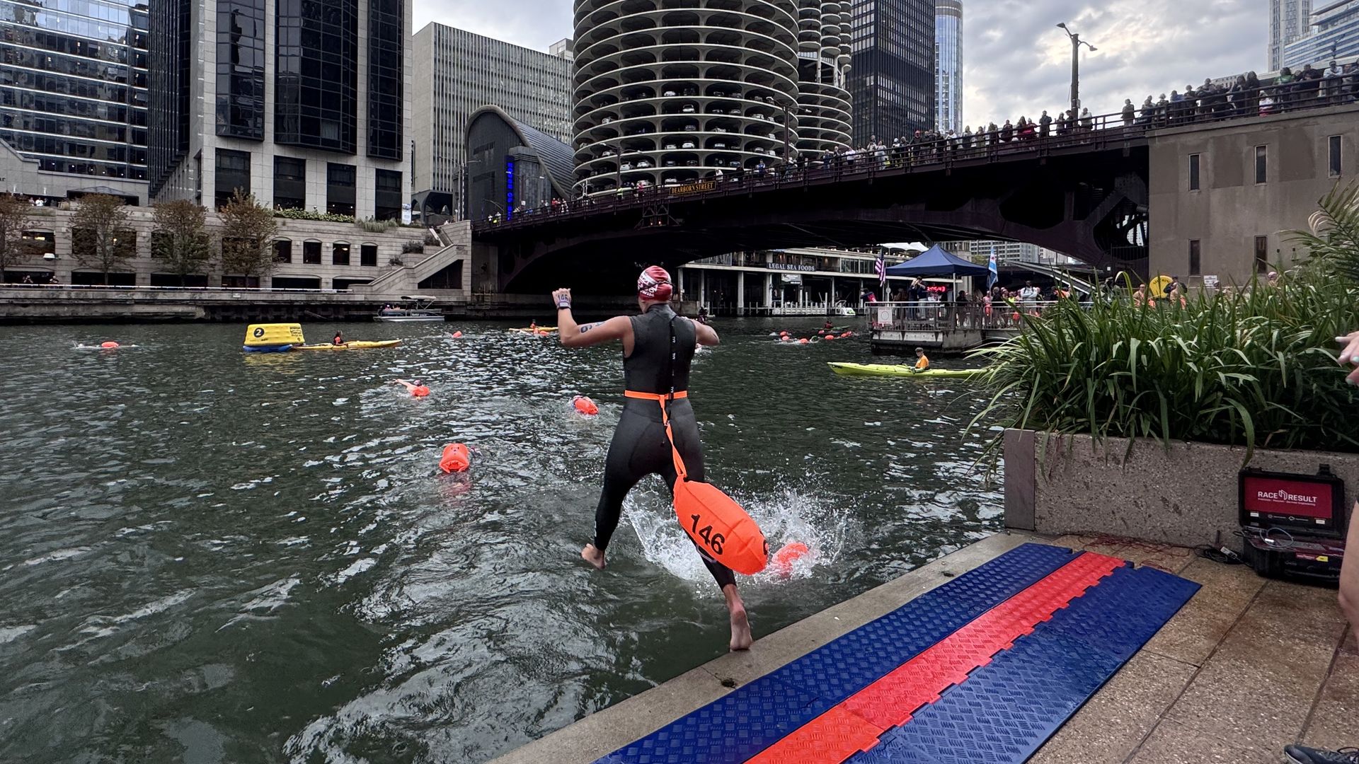 Triathlete in a black wetsuit leaps from a platform lined with blue and red mats into the Chicago River, carrying an orange buoy marked 146. A crowded bridge and tall skyscrapers loom overhead.