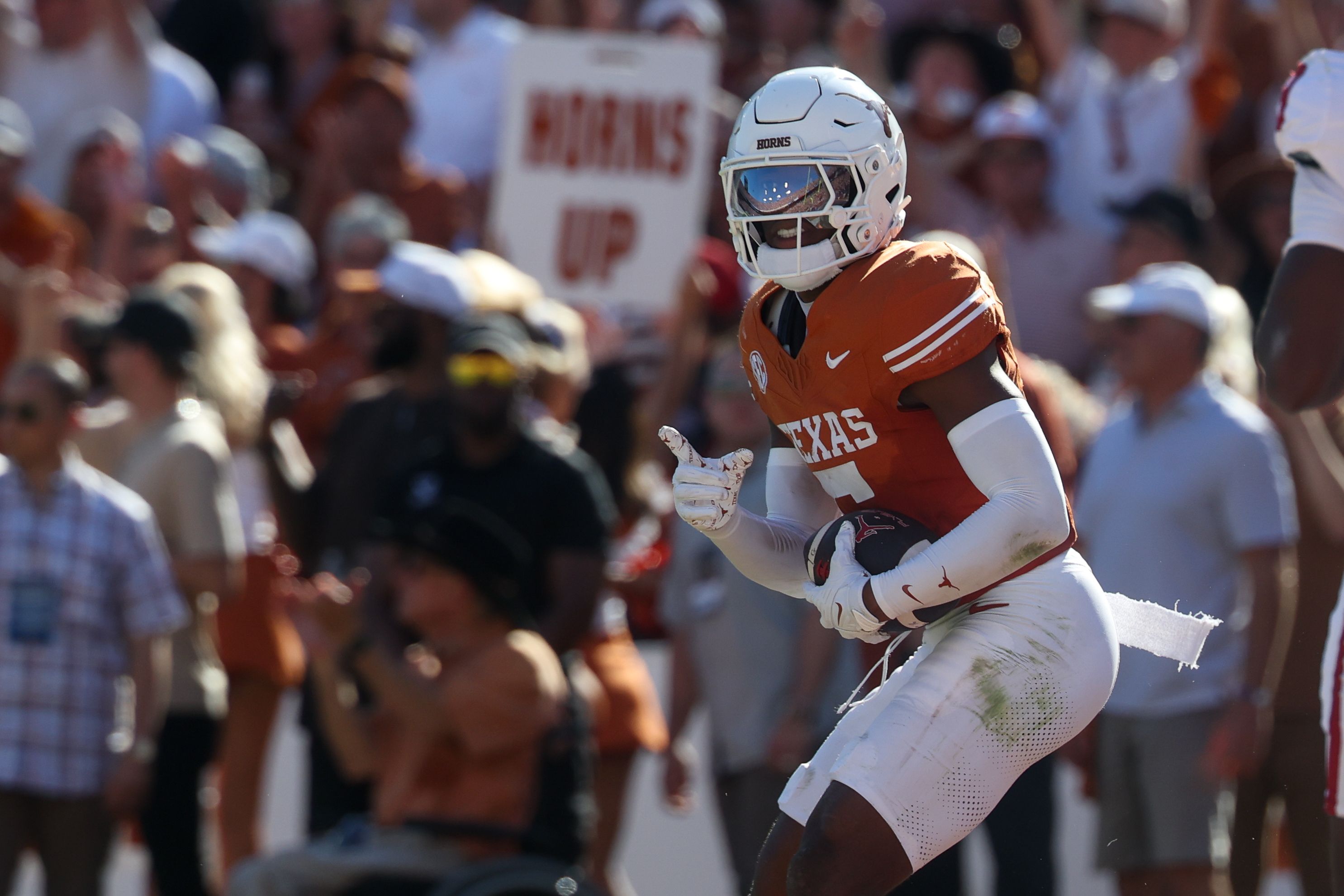 Texas Longhorns football player in an orange jersey and white helmet labeled HORNS runs with the ball during a game, crowd blurred in background.