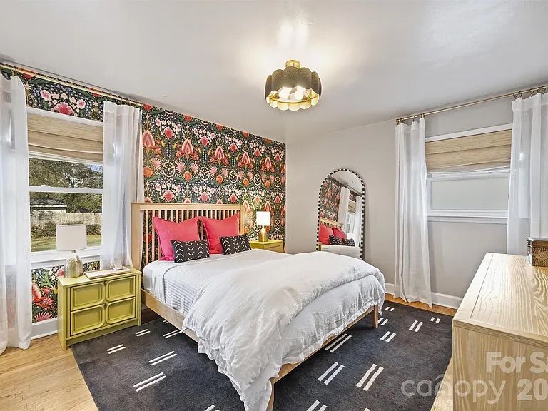 Bedroom with floral wallpaper wall, wooden bed with white bedding and pink and black pillows, green nightstands, large arched mirror, sheer white curtains, and modern light fixture.