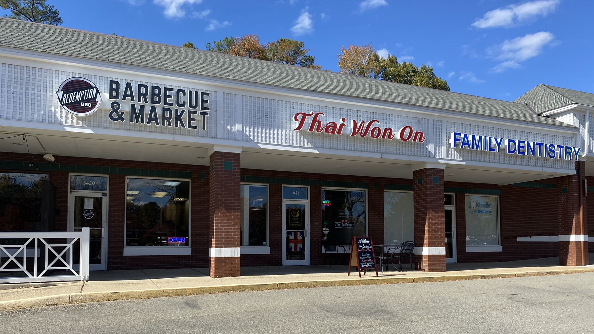 Strip-mall storefronts beneath a blue sky, signs read "BARBECUE & MARKET" with a circular "Redemption BBQ" logo, red "Thai Won On", and blue "FAMILY DENTISTRY". Brick columns, windows, and a chalkboard sign outside.