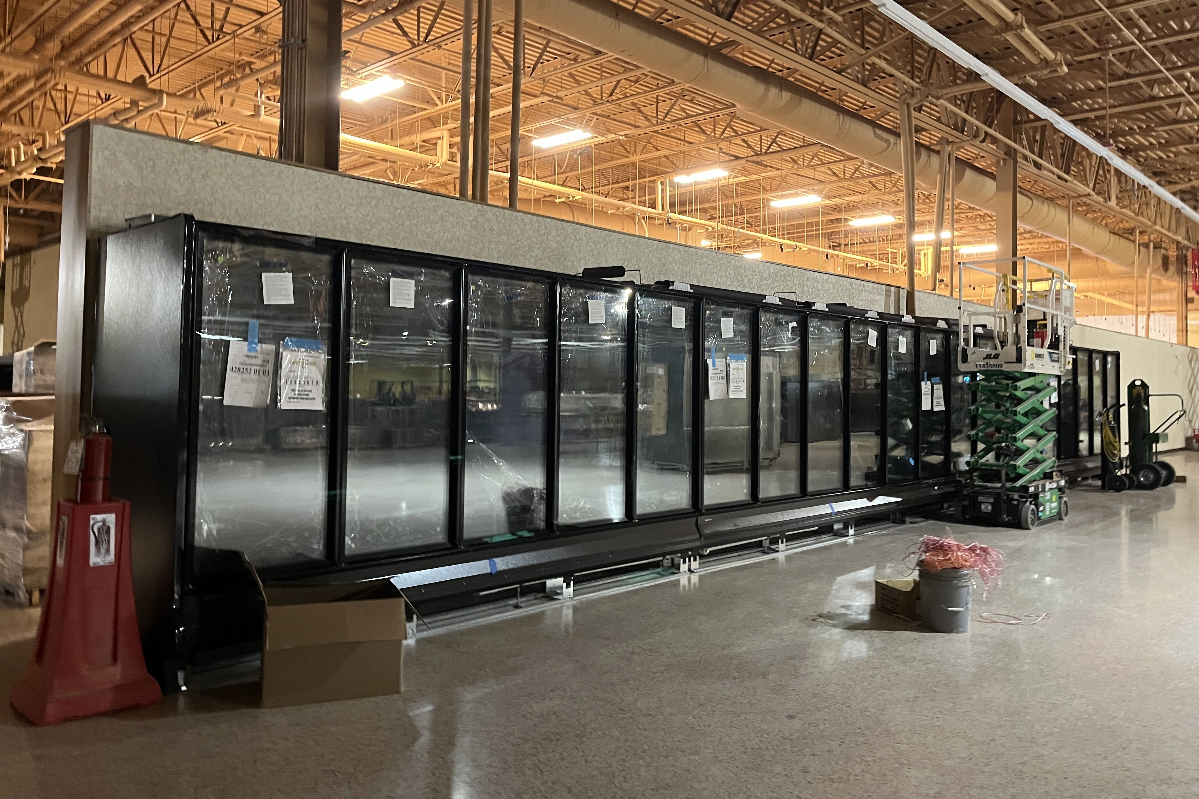 Inside a large industrial space under construction, a row of black-framed glass-front refrigerators sits against a gray wall, with overhead ducts, a raised scissor lift, and safety gear nearby.