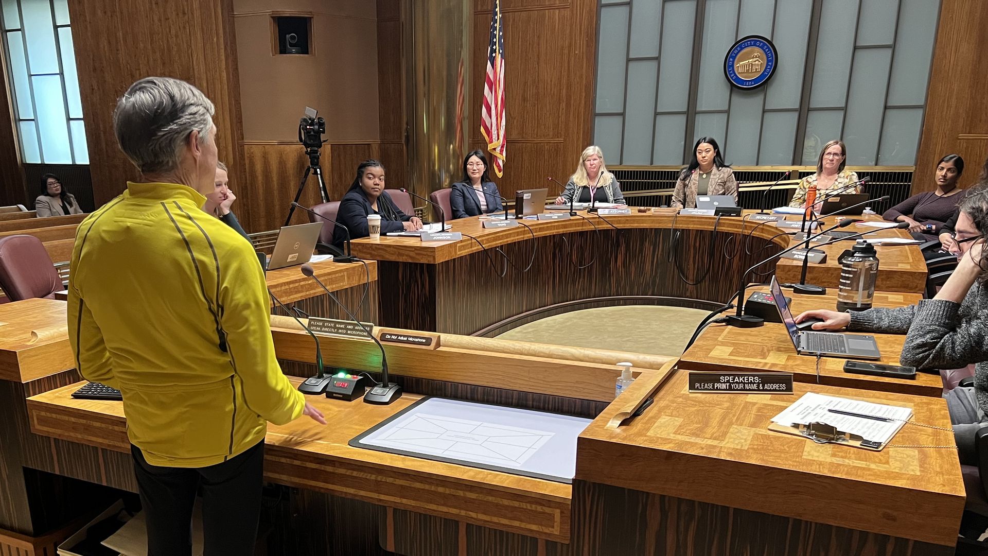 A man in a bright yellow shirt stands at a horseshoe-shaped dais and addresses a city council