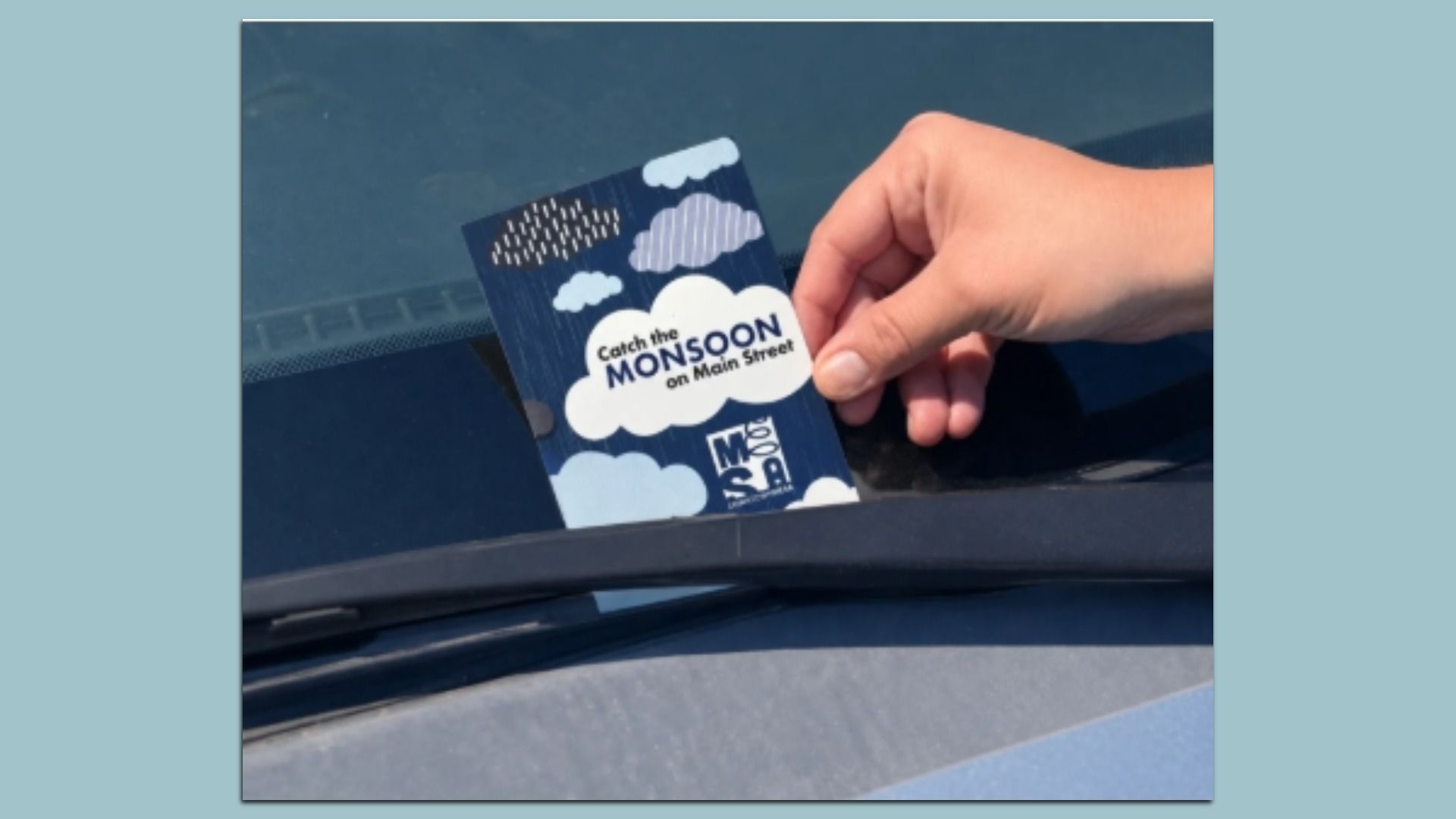 Close-up of a hand placing a blue promo card with white clouds and rain on a car window. The card reads, "Catch the MONSOON on Main Street."