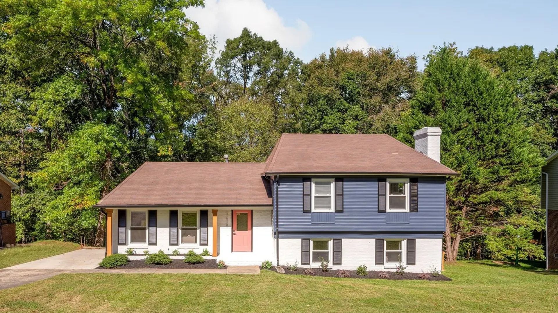Two-story house with blue and white exterior, brown roof, coral front door, black shutters, surrounded by green lawn and tall trees under clear sky.
