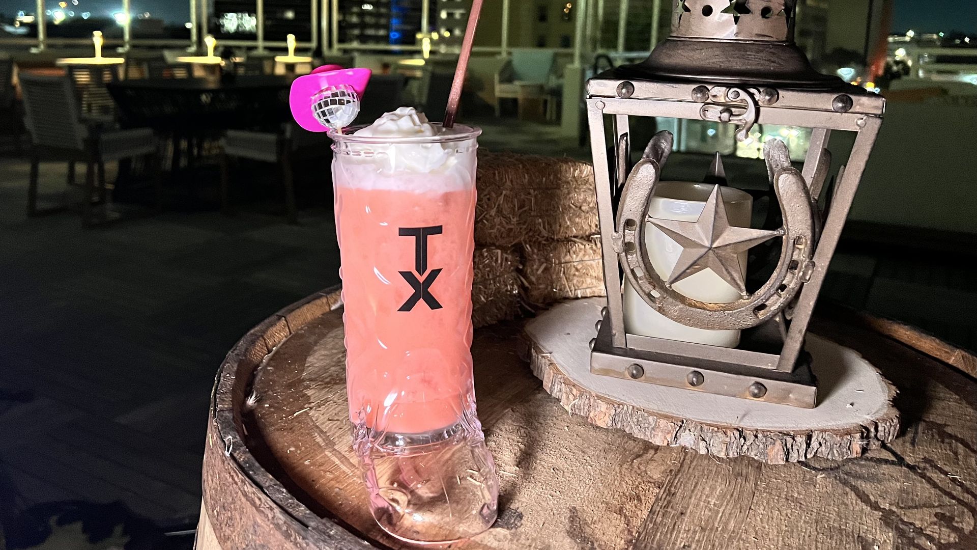 Night rooftop scene with a pink cocktail in a boot-shaped glass with TX logo, topped with whipped cream and a pink umbrella, next to a rustic metal lantern with a star and horseshoe design.
