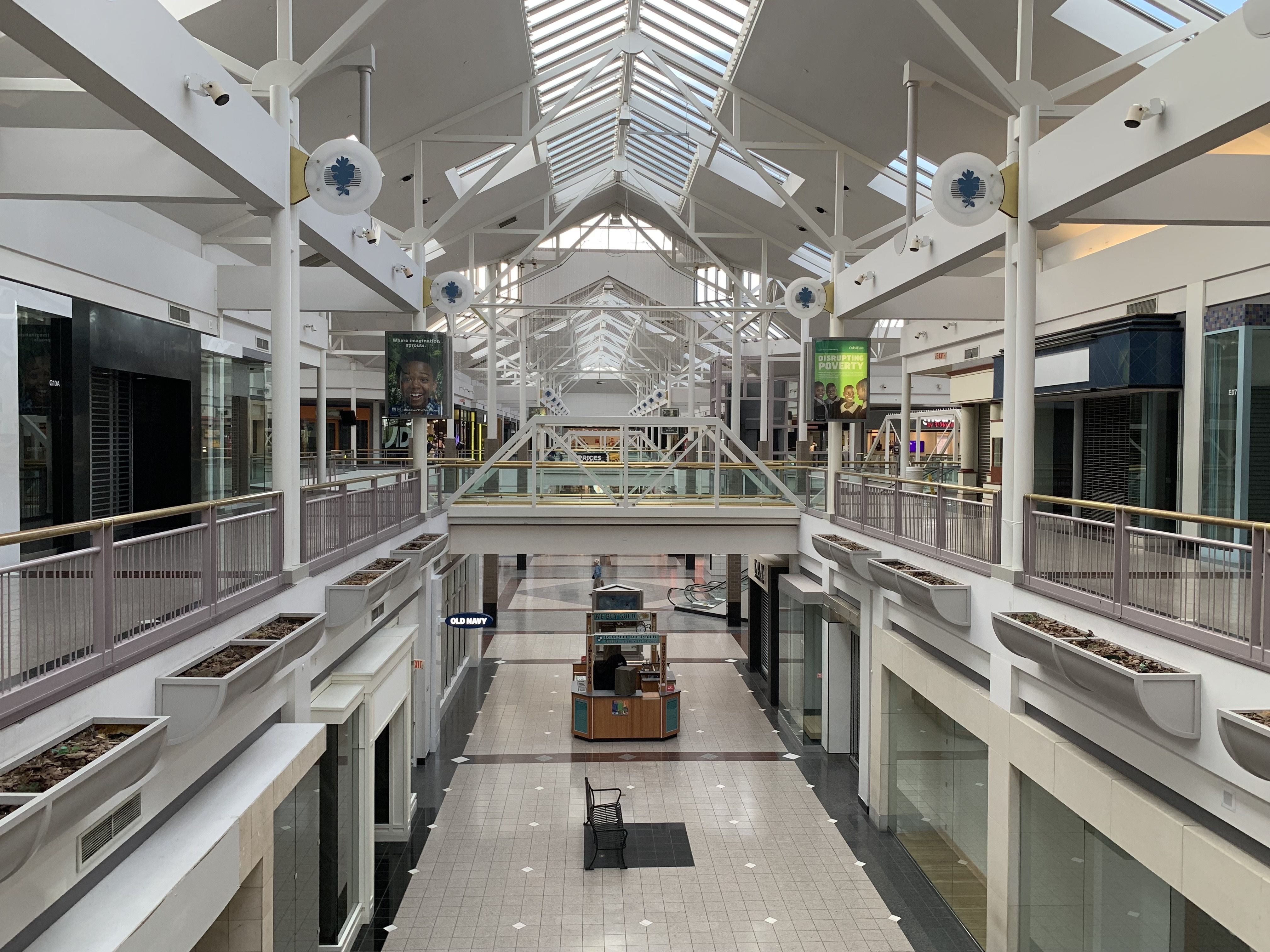 Bright white mall atrium with exposed steel trusses and glass skylights. Central corridor with rails, benches, banners, and planters; round blue-leaf logo fans along rails; storefronts on both sides.