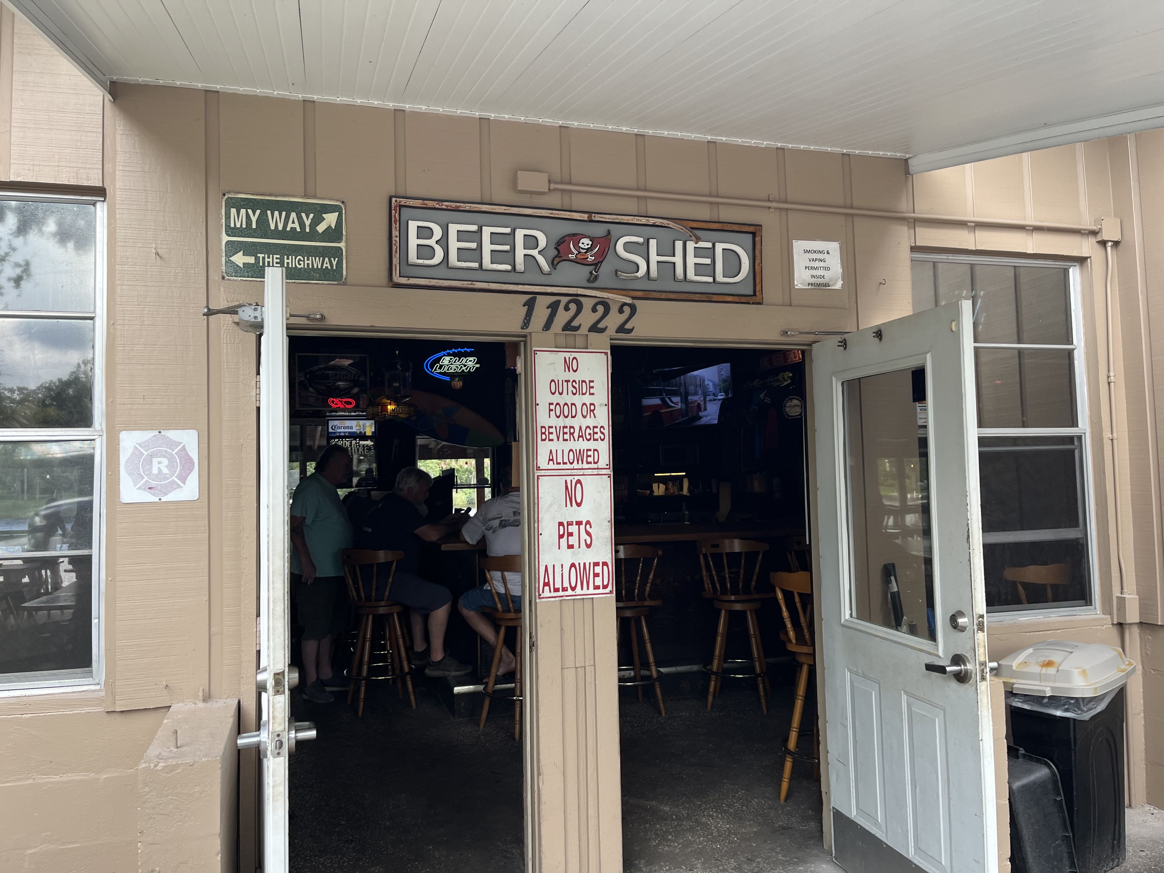 Entrance to the 'BEER SHED' bar with a pirate flag sign and the number 11222 above the door; a green 'MY WAY' sign to the left. Inside, patrons sit at tables; a white sign prohibits outside food or beverages.