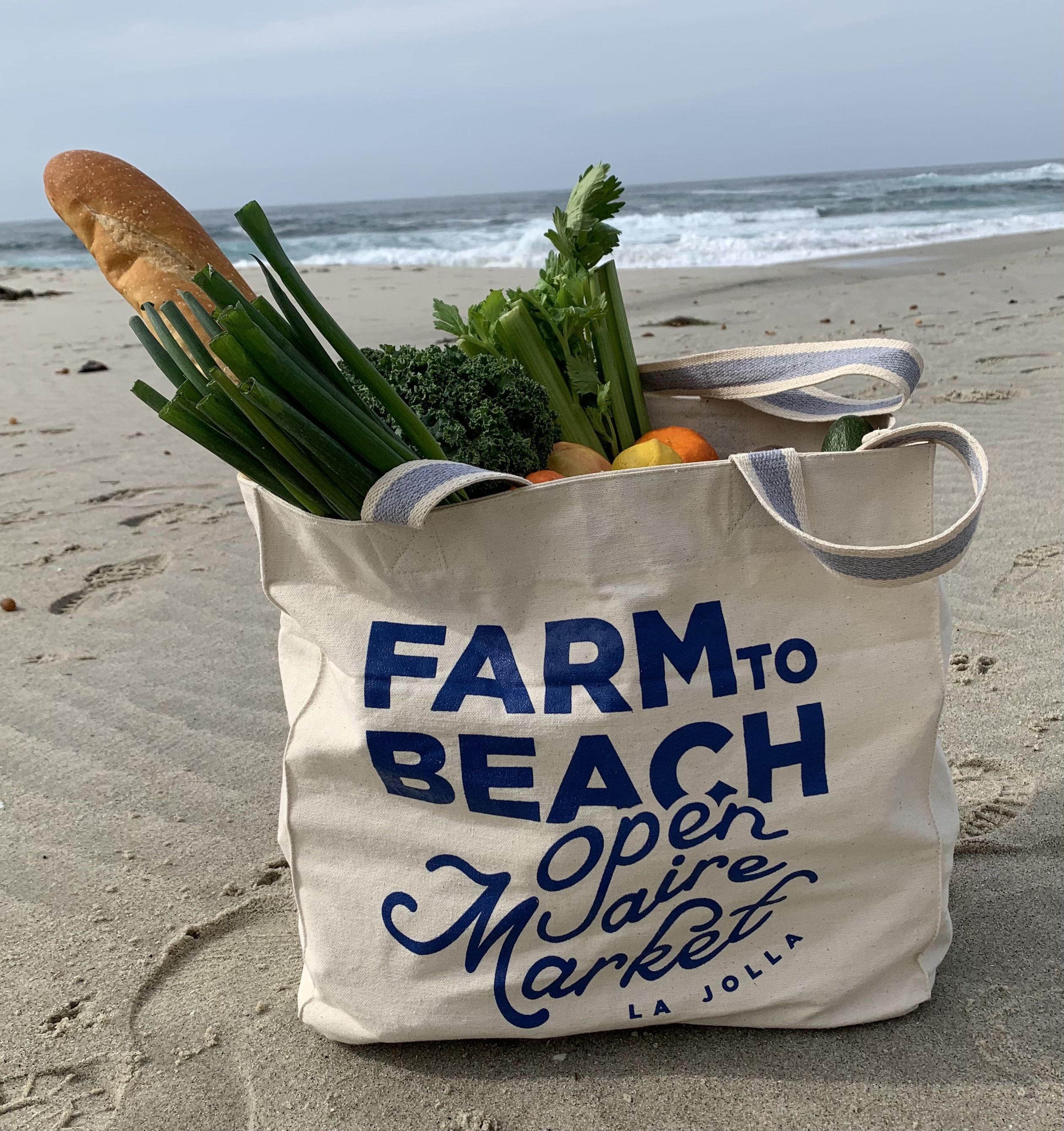 A tote bag that says "Farm to Beach open aire market La Jolla" with fresh produce sits on a sandy beach.