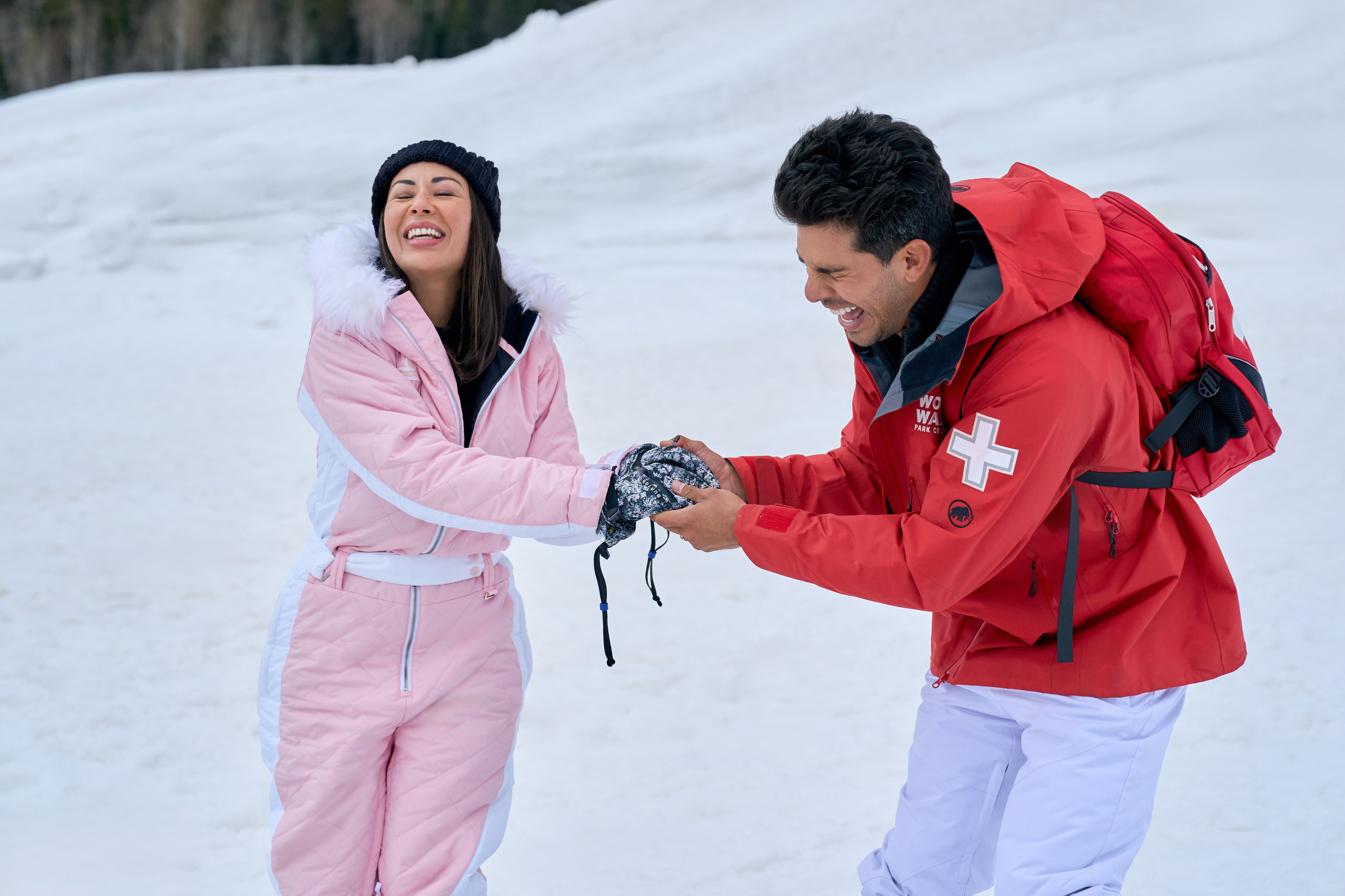 A man in a red jacket with a white cross and a woman in a pink snow suit laugh while holding a black and white winter hat together on a snowy slope.