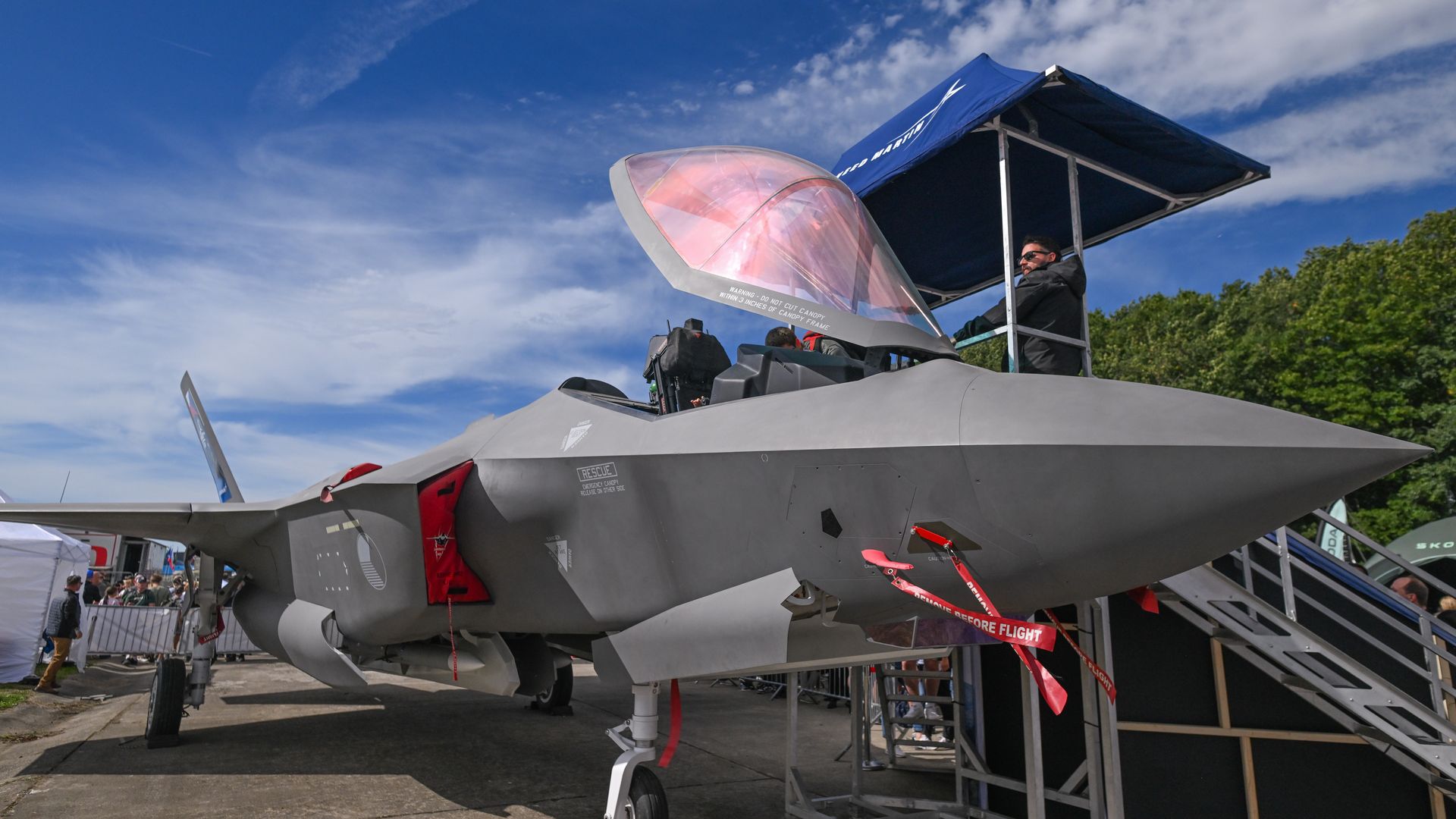 Side view of a gray military jet on display, cockpit canopy, red remove-before-flight ribbons, blue canopy shelter overhead, with spectators and barriers under a blue sky.