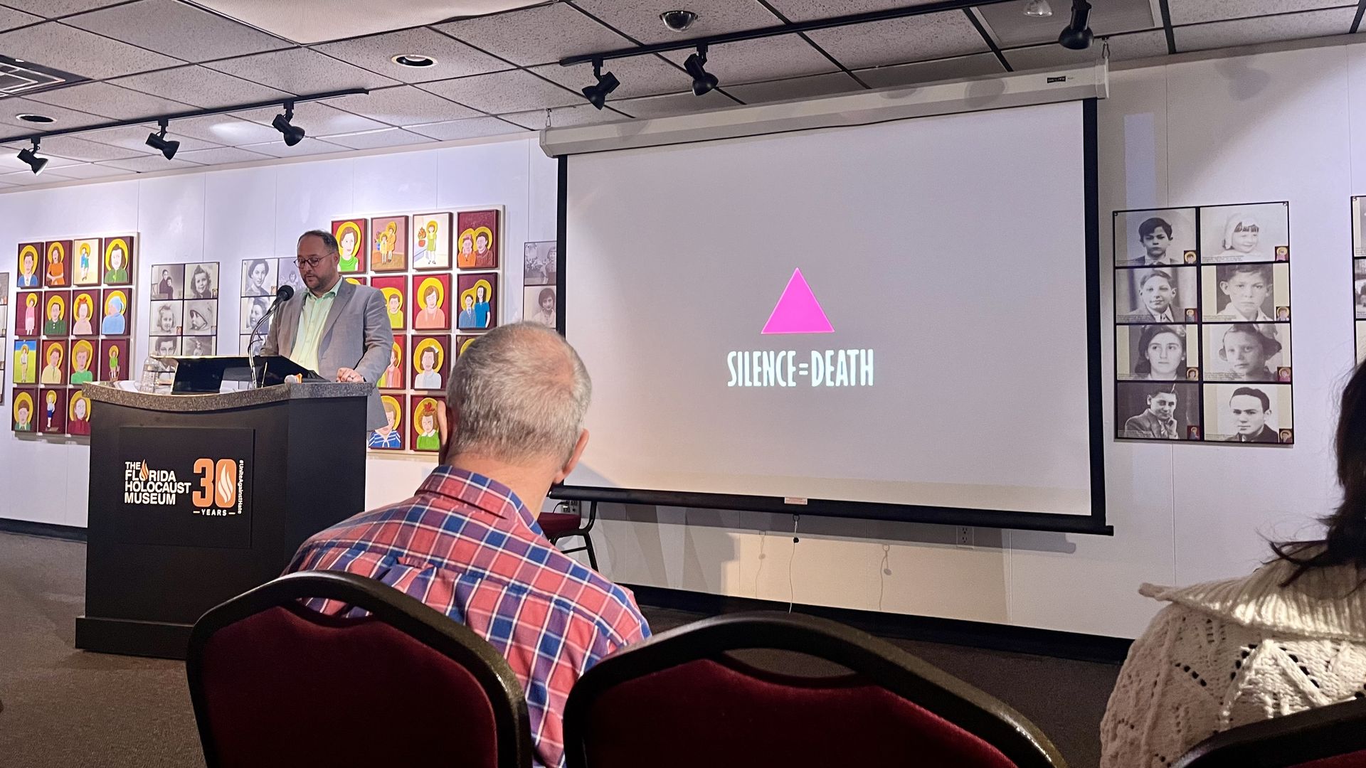 A man stands at a lectern with a sign for the Florida Holocaust Museum. To his right is a projector screen showing a bright pink triangle and beneath it, "SILENCE = DEATH."