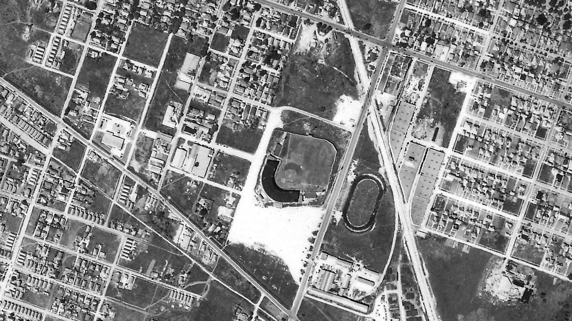 Grayscale aerial view of a city: a dense street grid with several parks, and a large oval track inside a park near the center-right, surrounded by houses, roads, and open spaces.