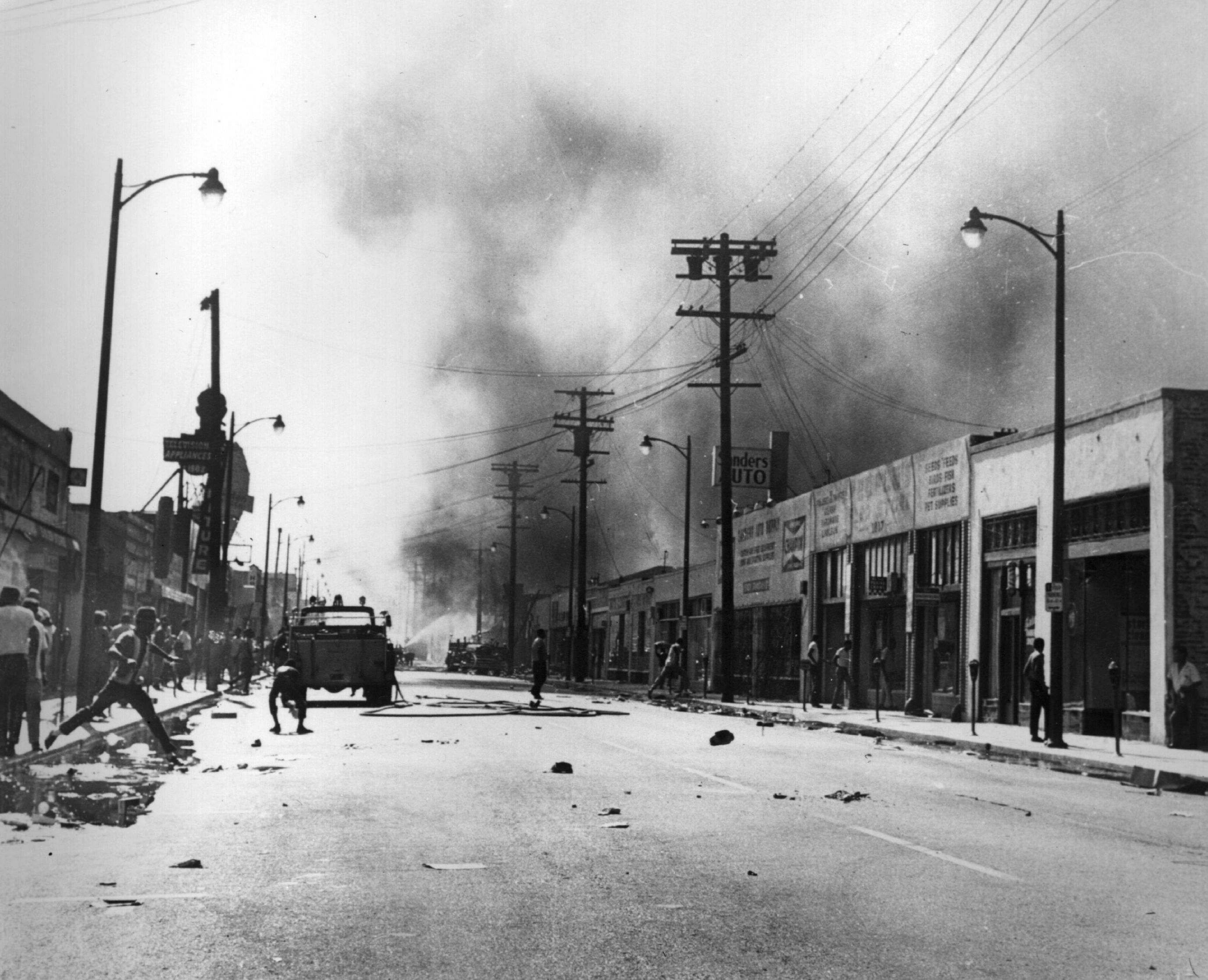 Black and white photo of a city street with smoke rising in the distance, people scattered along sidewalks, a fire truck in the middle, and debris on the road.