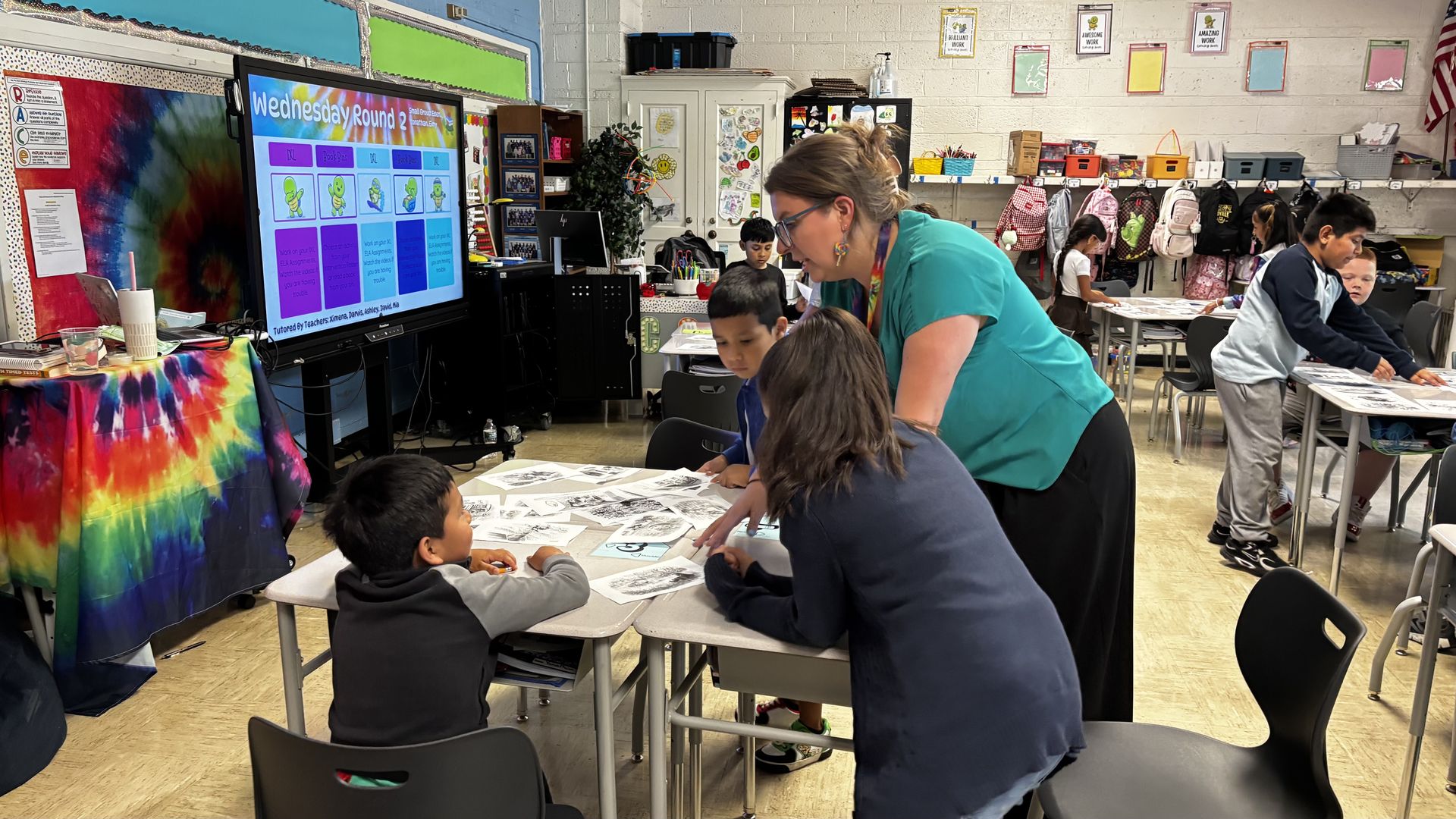 Teacher in teal assists students at desks in a colorful classroom. A large screen reads "Wednesday Round 2" with colorful tiles; backpacks on the wall, tie-dye cloth, and posters decorate the room.