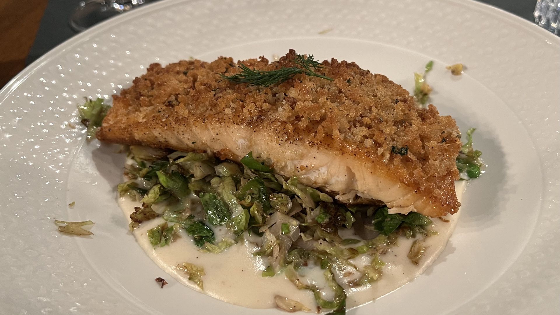 Plate with a crispy crusted salmon fillet on a bed of sautéed greens and creamy white sauce, garnished with a small sprig of fresh dill on top.