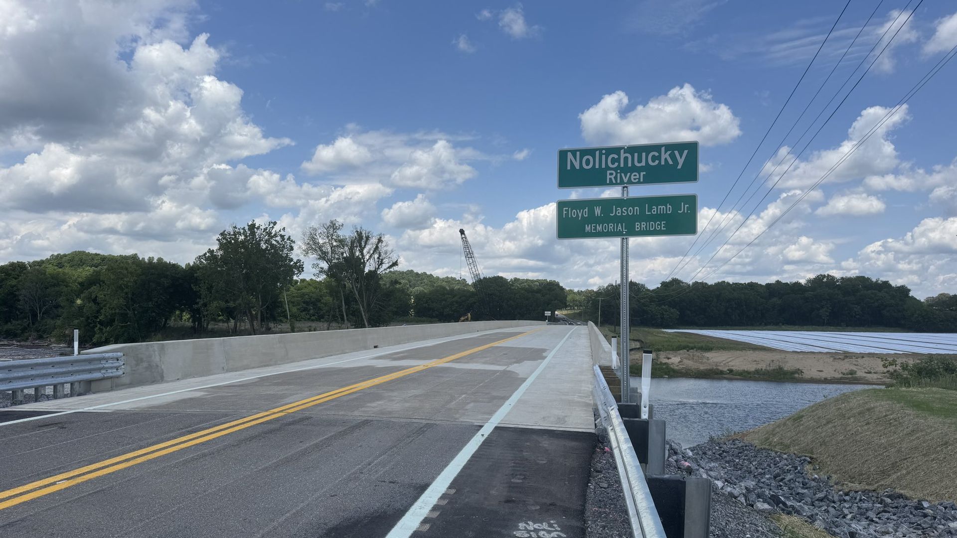 Bridge over the Nolichucky River on a sunny day. Green signs read "Nolichucky River" and "Memorial Bridge." Blue sky with white clouds, trees on the left, power lines on the right.