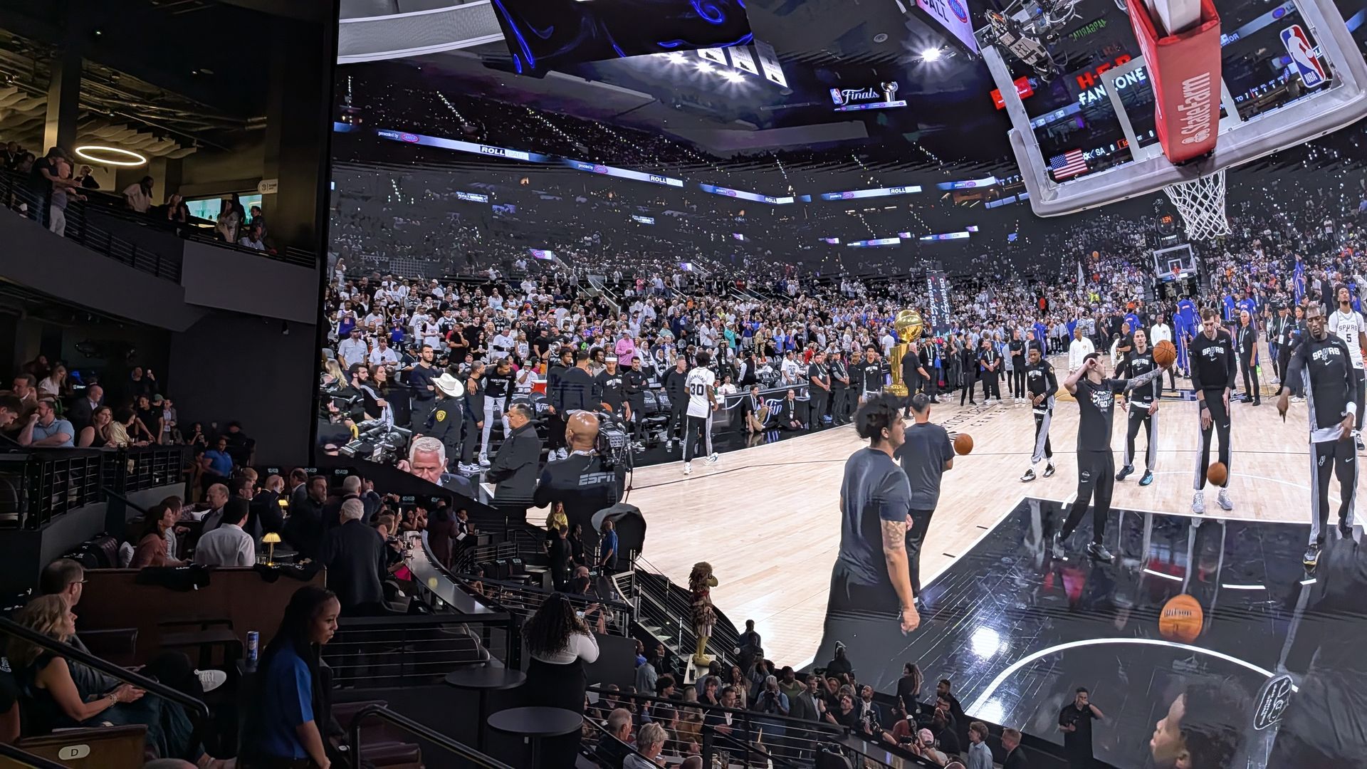 An LED screen dome showing a packed basketball arena, fans fill the stands and court side areas. A hoop dominates the right foreground while players in black uniforms gather nearby and a trophy sits on the court.