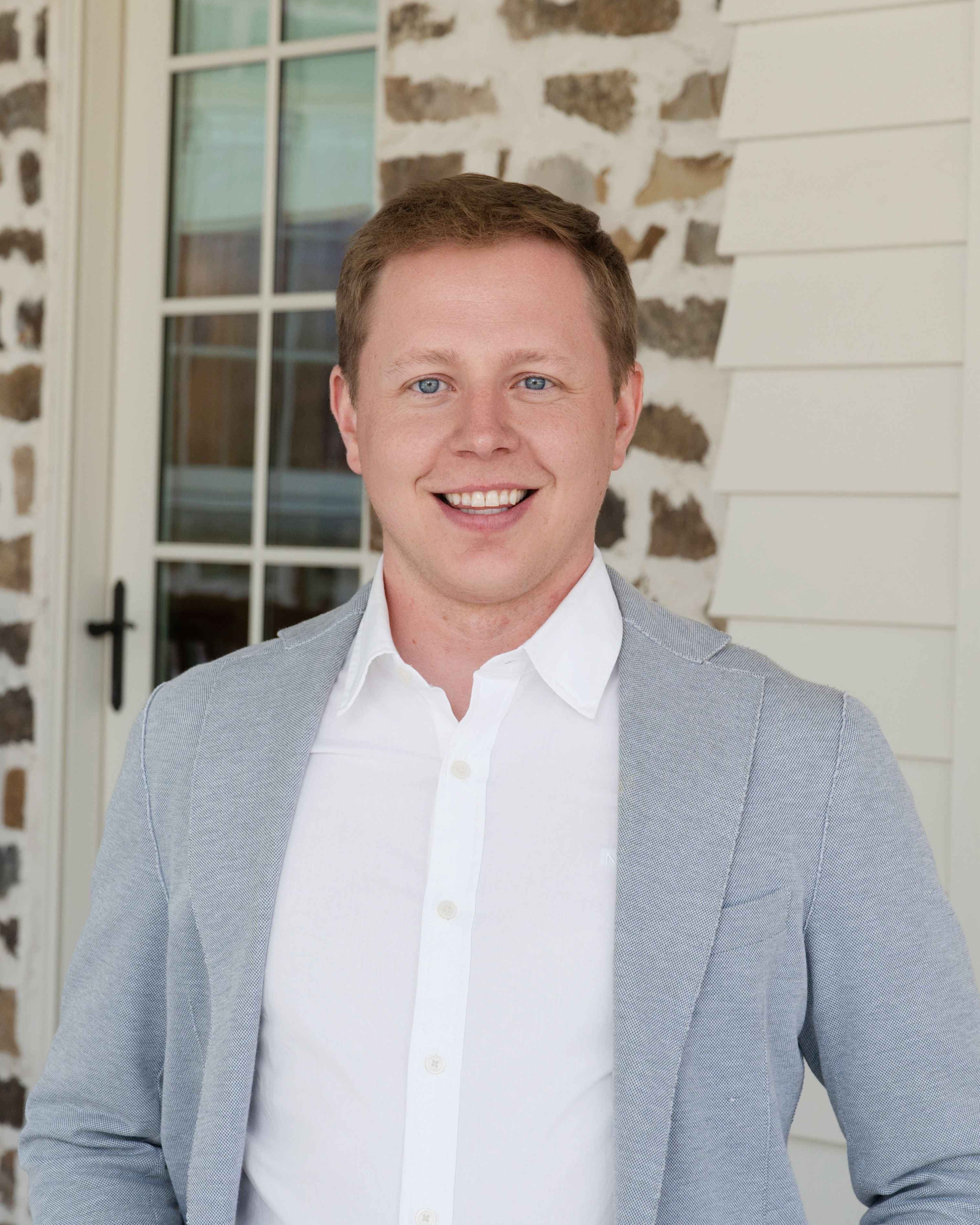 Smiling man with short light brown hair wearing a white shirt and light gray blazer, standing in front of a stone wall and windowed door.