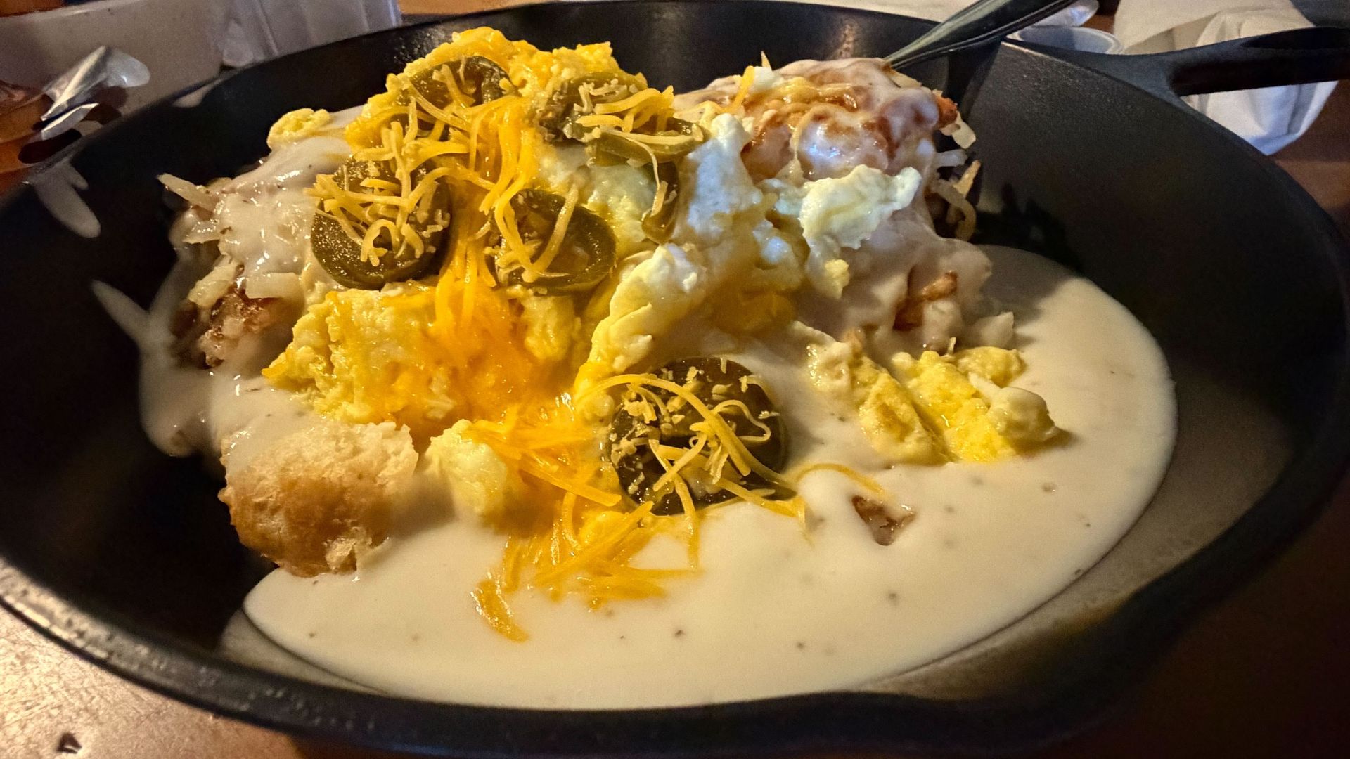 Breakfast skillet with scrambled eggs, shredded cheddar, jalapeño slices, biscuits, sausage, and white gravy served in a black cast iron pan.