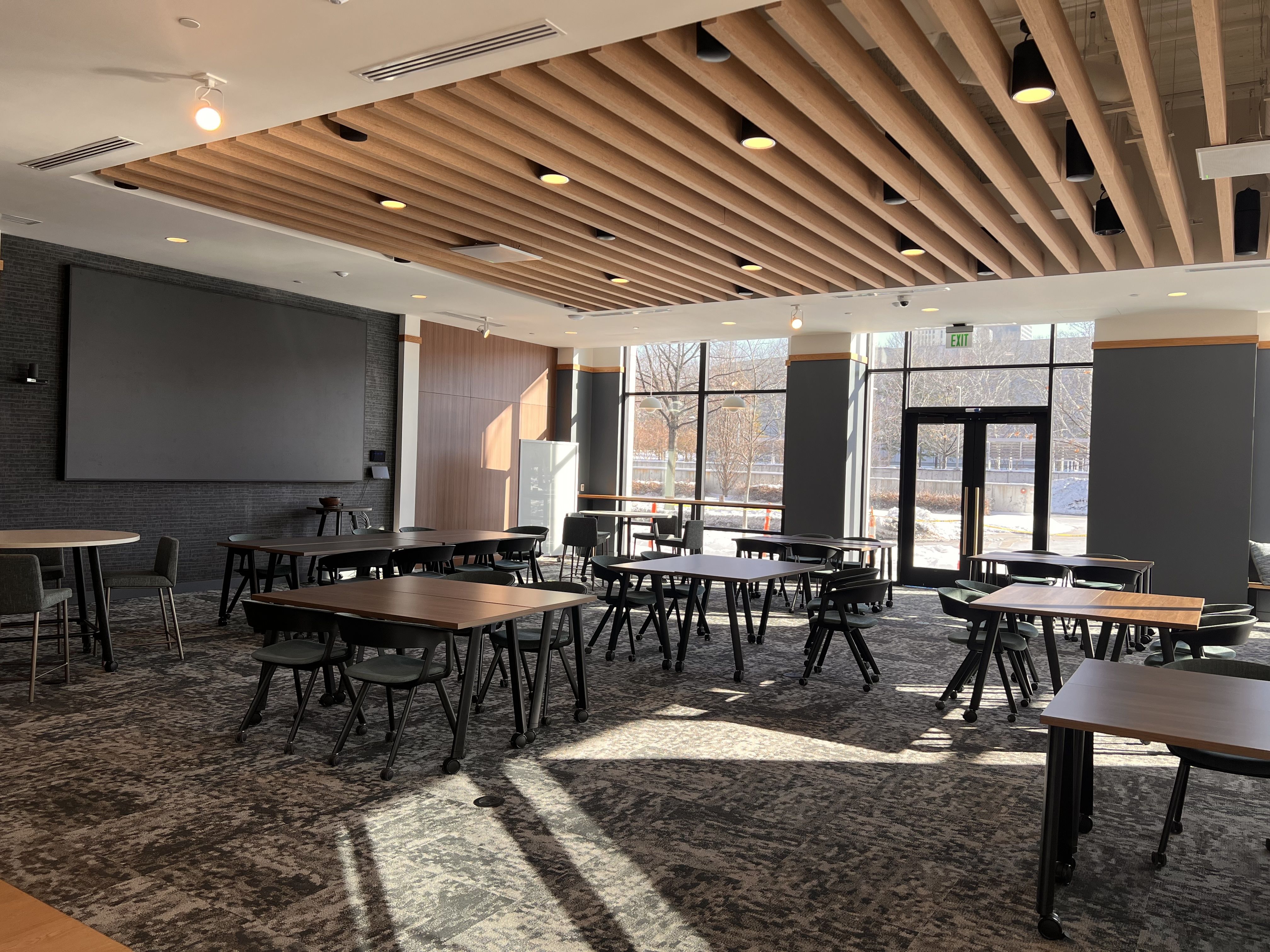Sunlit modern meeting room with wooden tables and dark chairs on patterned carpet, large windows letting in natural light, wooden beam ceiling, and a large blank screen on a dark wall.