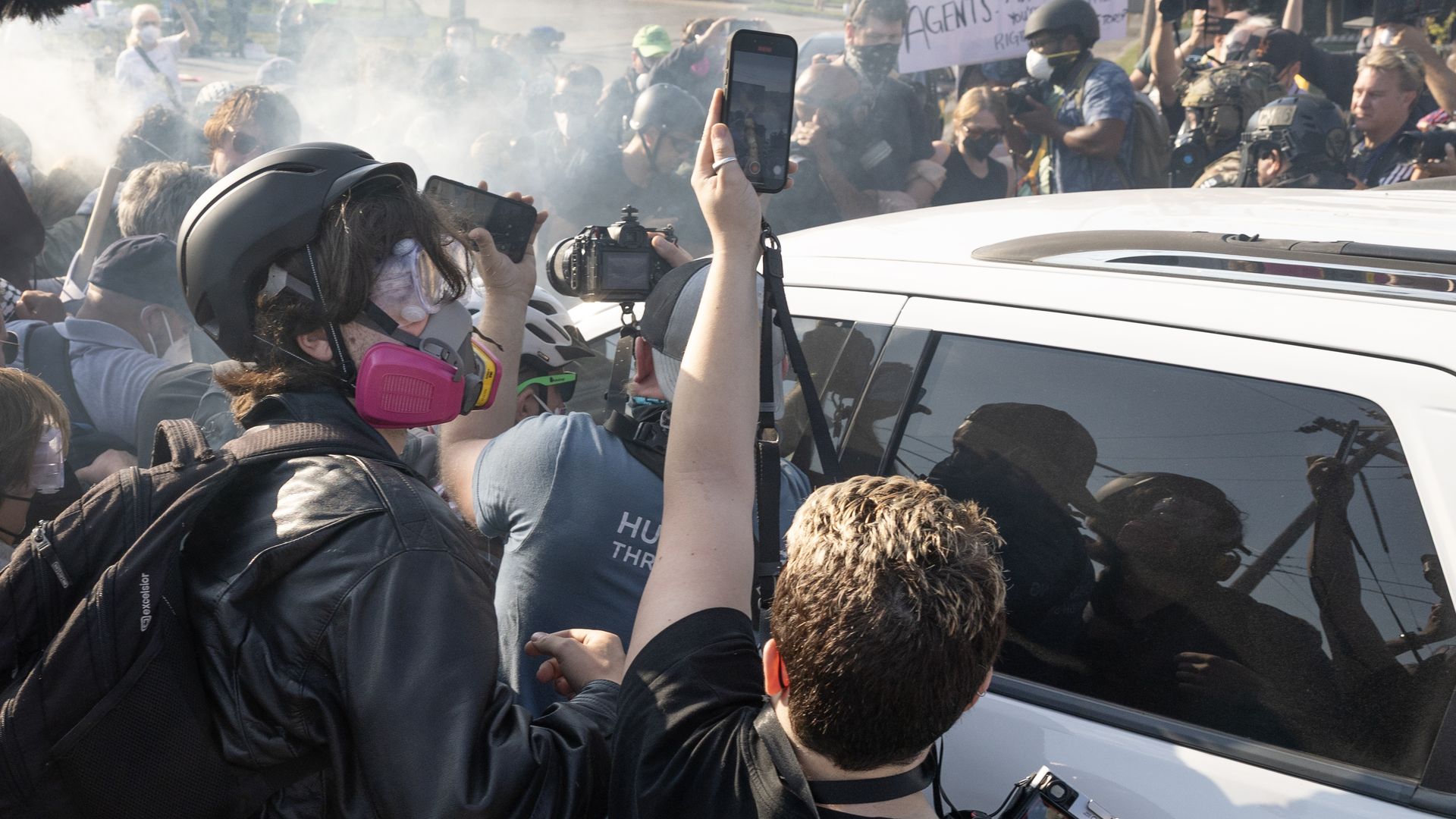 Photo of protesters surrounding a car as smoke rises in the air. 