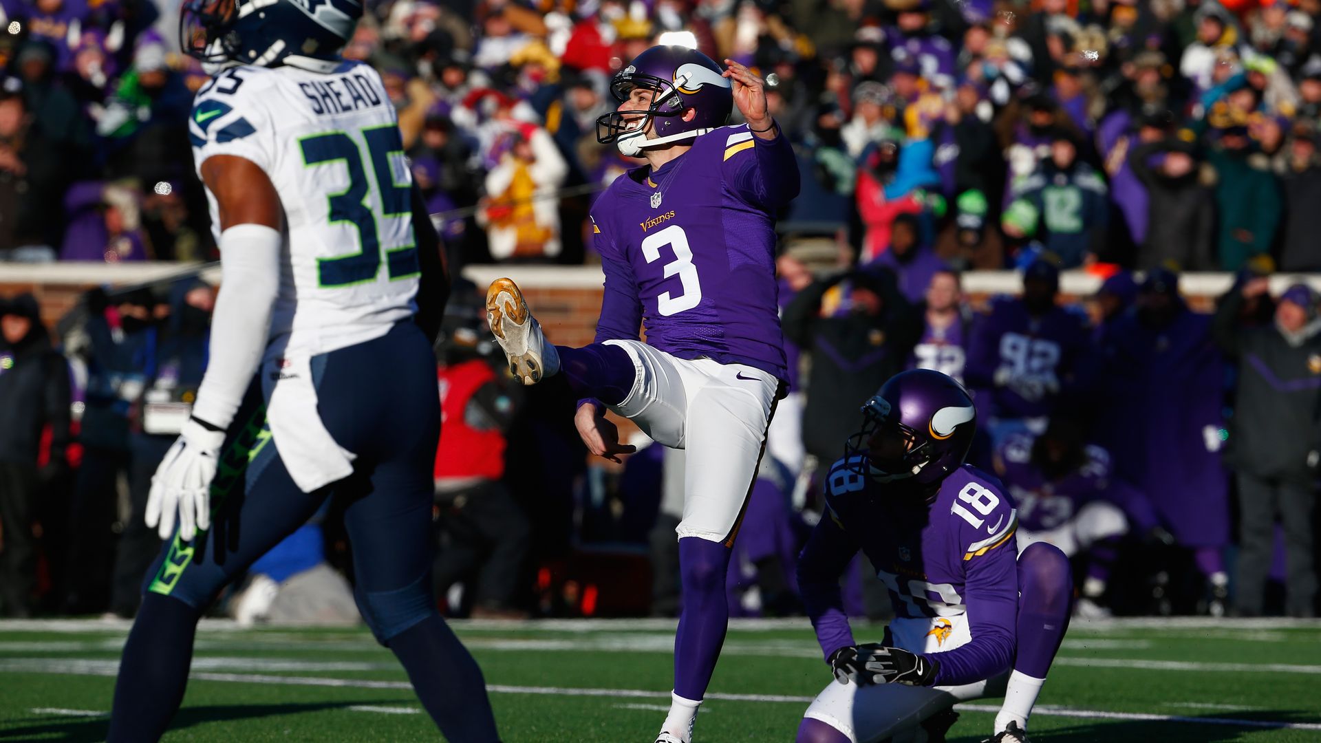 Minnesota Vikings player kicking a football held by a teammate on a green field, with a Seattle Seahawks player in white and blue nearby and a crowd in the background.