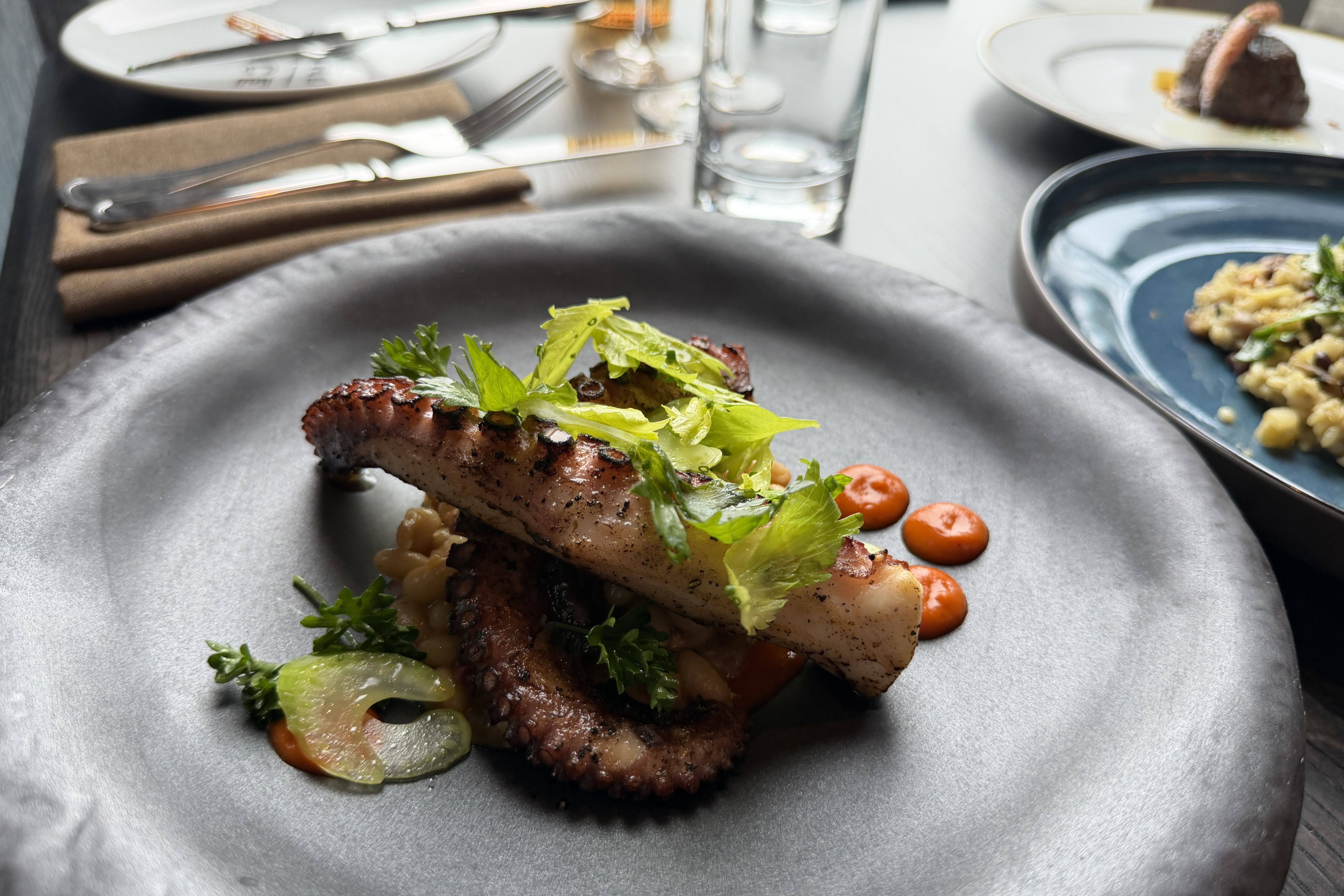 Close-up of a plated dish: grilled protein topped with greens on a bed of grains, with small orange sauce drops on a dark plate; blurred table, cutlery, glassware, and other dishes in the background.