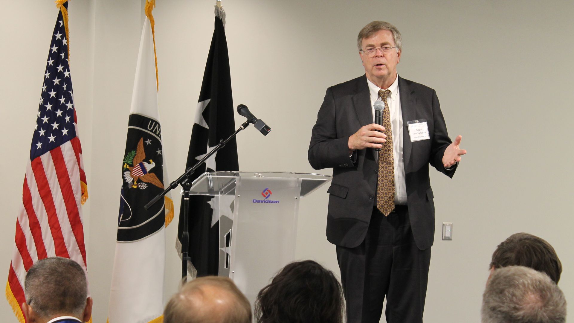 Man in a dark suit delivers a speech at a podium with a microphone, as the American flag and several other flags stand behind him at a formal event.