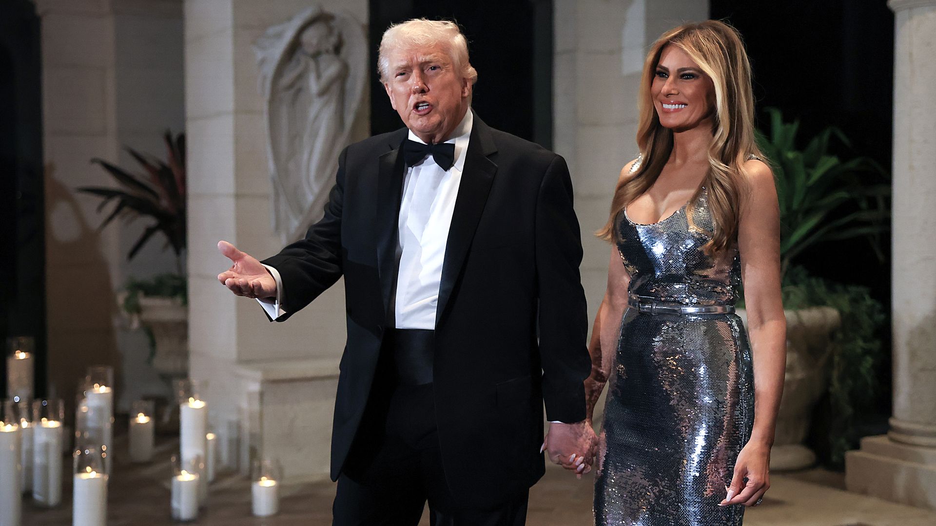 President Trump, in black tuxedo with bow tie speaking, holding hands with first lady Melania Trump in sleeveless, silver sequined dress standing in a stone building with candles on floor.
