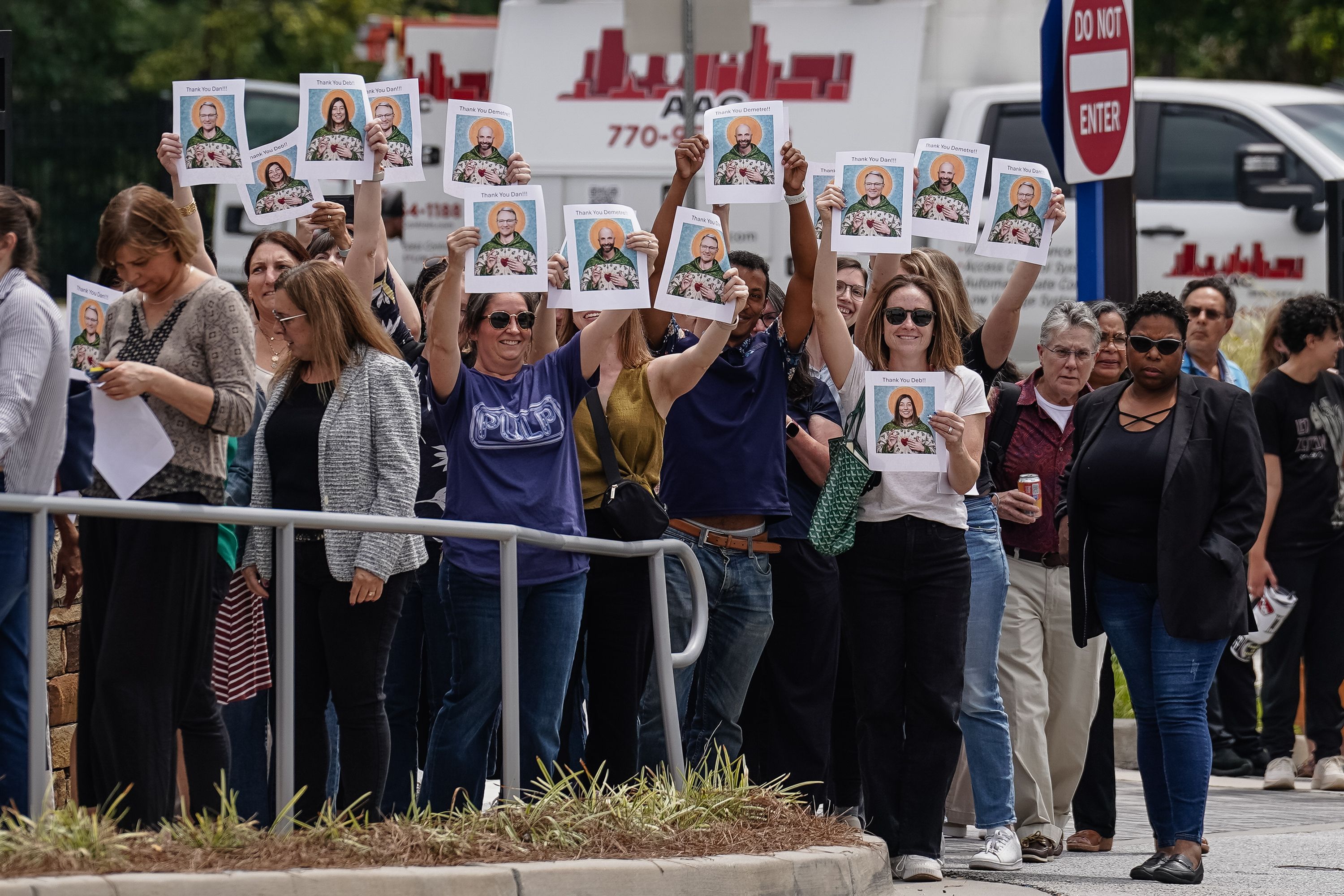 A diverse group of people standing outside, many holding up printed photos of two different smiling individuals with "Thank You" messages, near a "Do Not Enter" sign and vehicles.